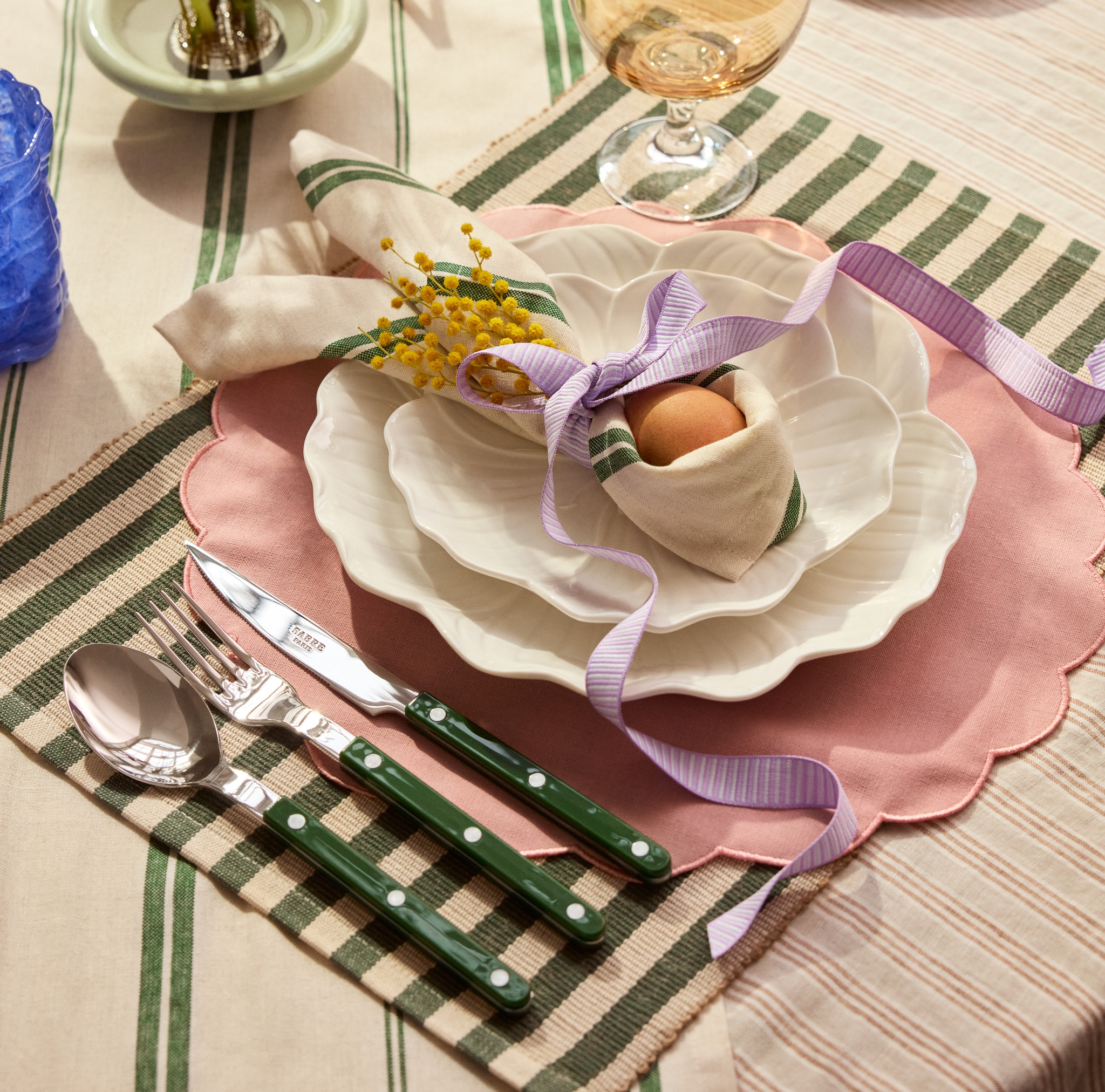 En vårdekket bord med bølgete tallerkener, en rosa bordbrikke, en grønnstripet serviett med et egg og gul mimosa bundet med et lilla bånd, bestikk med grønne håndtak og lilla blomster