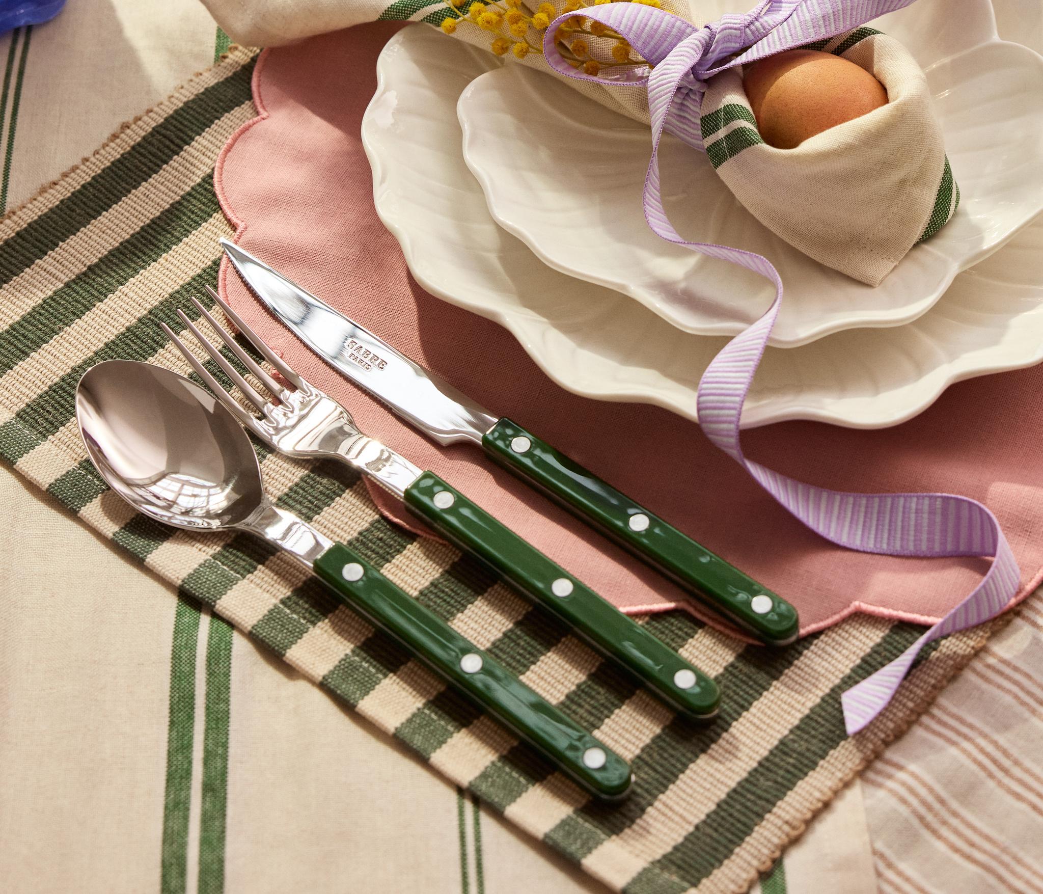 Keväinen kattaus, jossa on vaaleanpunainen aaltoreunainen pöytätabletti, valkoiset lautaset, pupunkorvien muotoon sidottu servetti, jonka sisällä on muna ja mimosankukkia, sekä vihreävartiset aterimet