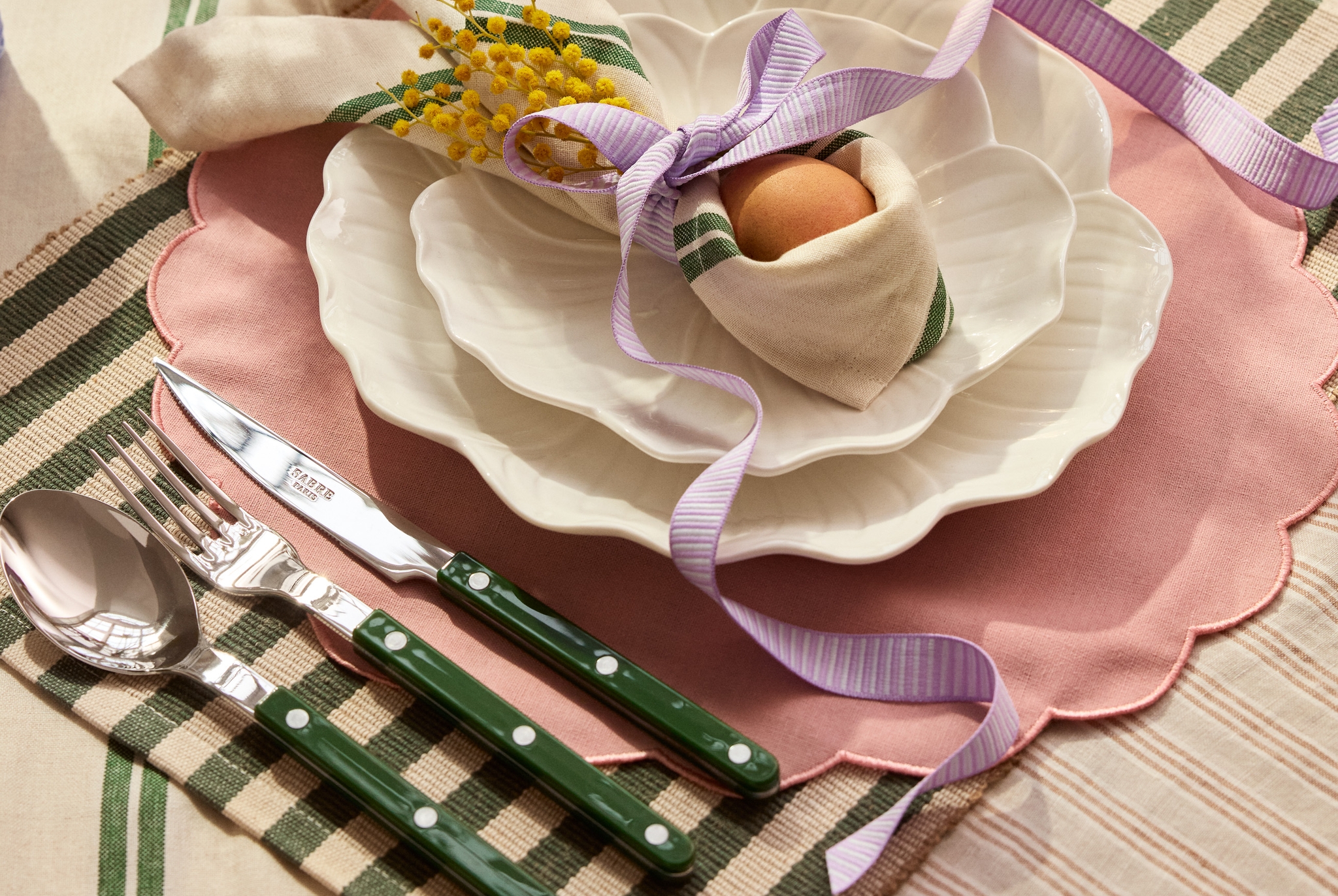 En festlig vårdukning med vita vågkantade tallrikar, gröna bestick, ett ägg inlindat i en servett och färgglada blommor