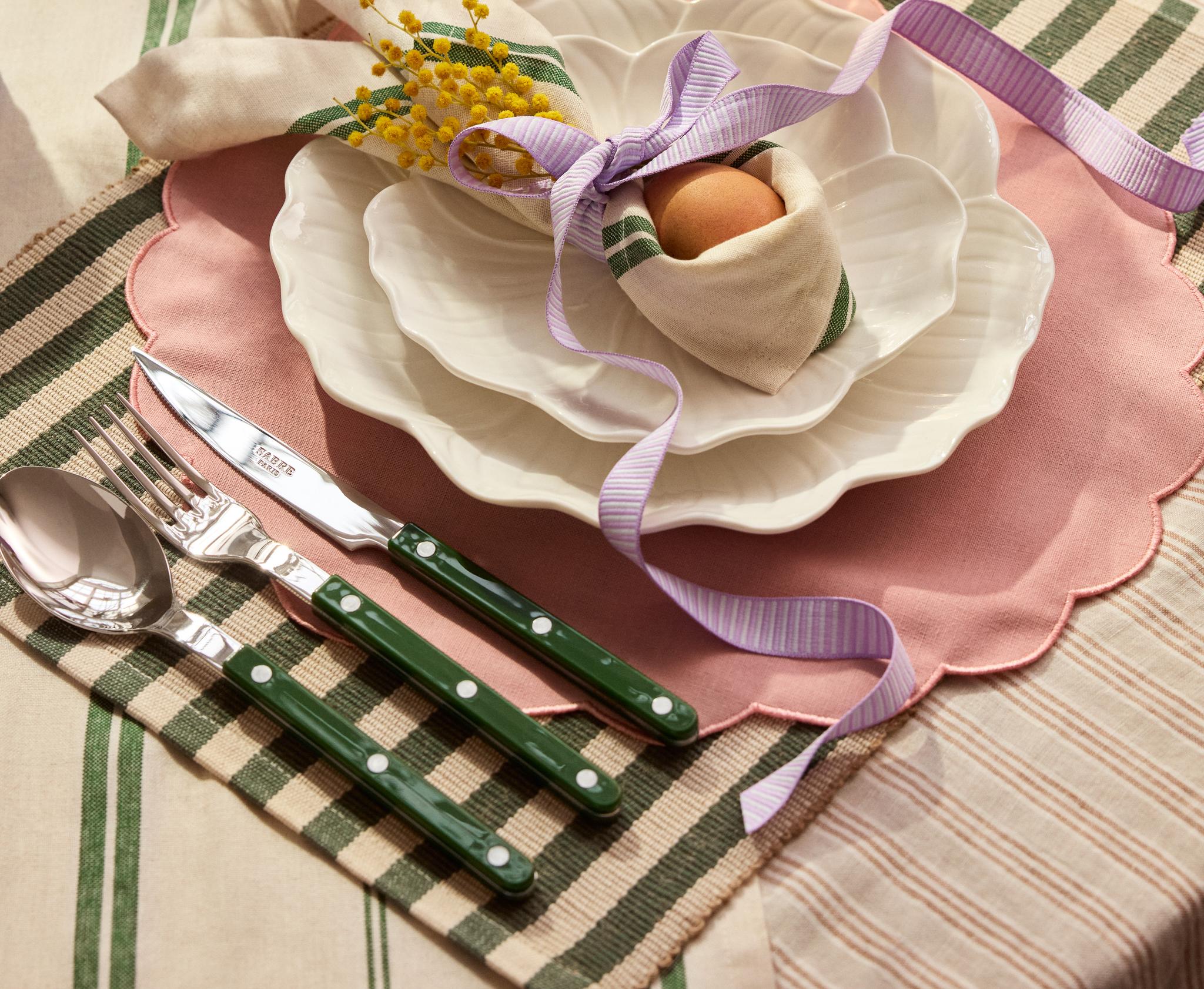 En vårdekket bord med bølgete tallerkener, en rosa bordbrikke, en grønnstripet serviett med et egg og gul mimosa bundet med et lilla bånd, bestikk med grønne håndtak og lilla blomster
