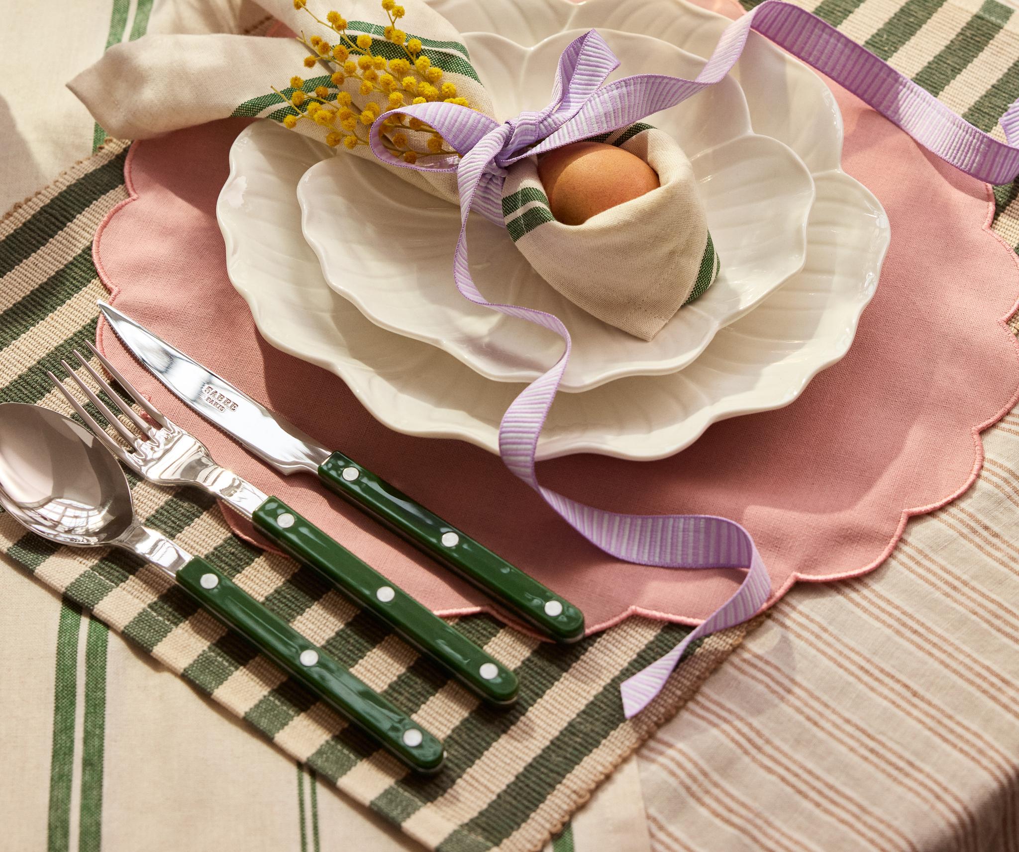 En festlig vårdukning med vita vågkantade tallrikar, gröna bestick, ett ägg inlindat i en servett och färgglada blommor