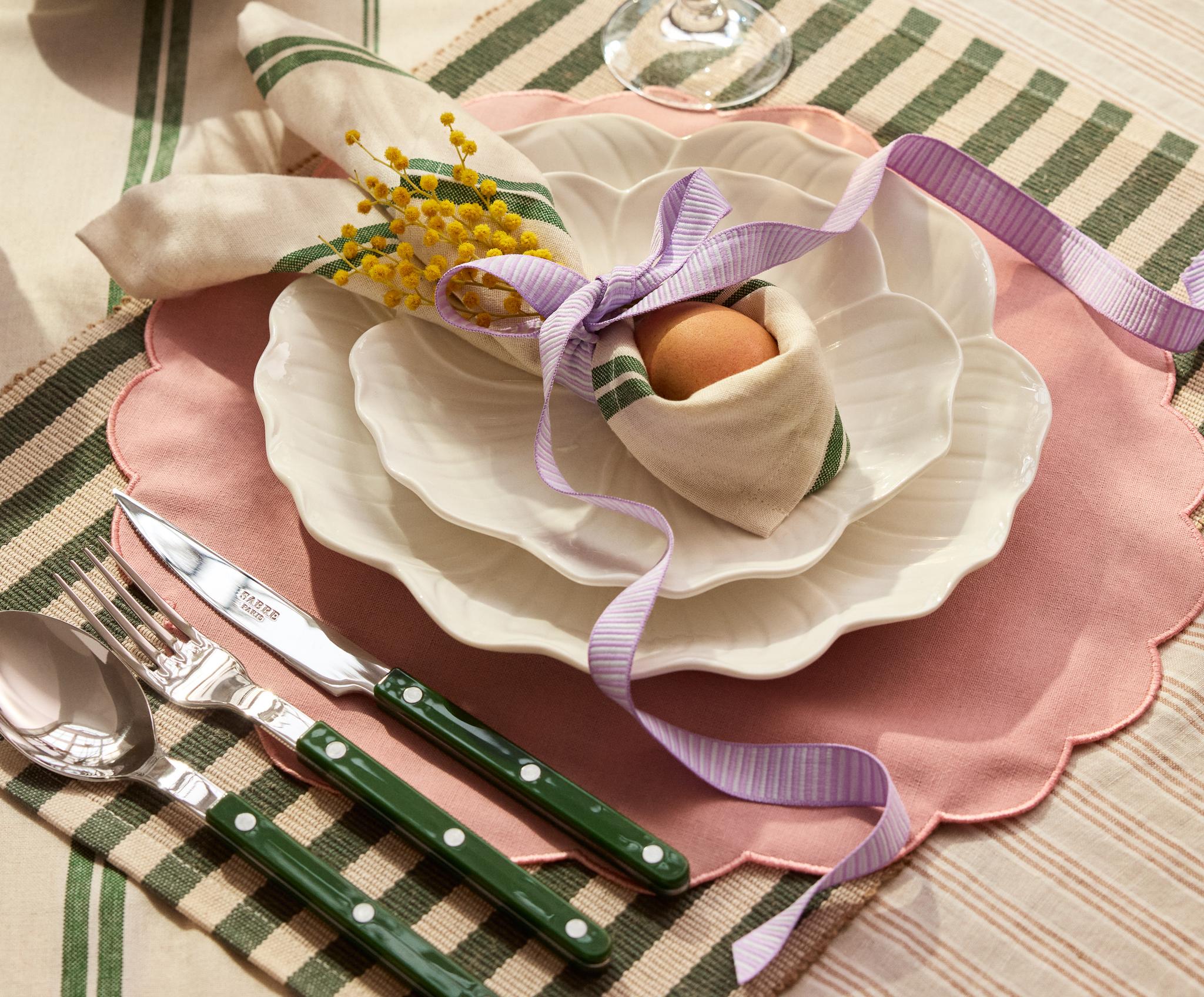 Keväinen kattaus, jossa on valkoiset aaltoreunaiset lautaset vaaleanpunaisella pöytätabletilla, raidallinen servetti, jossa on keltaista mimosa-kukkaa ja muna sidottuna violetilla nauhalla, sekä vihreävartiset aterimet