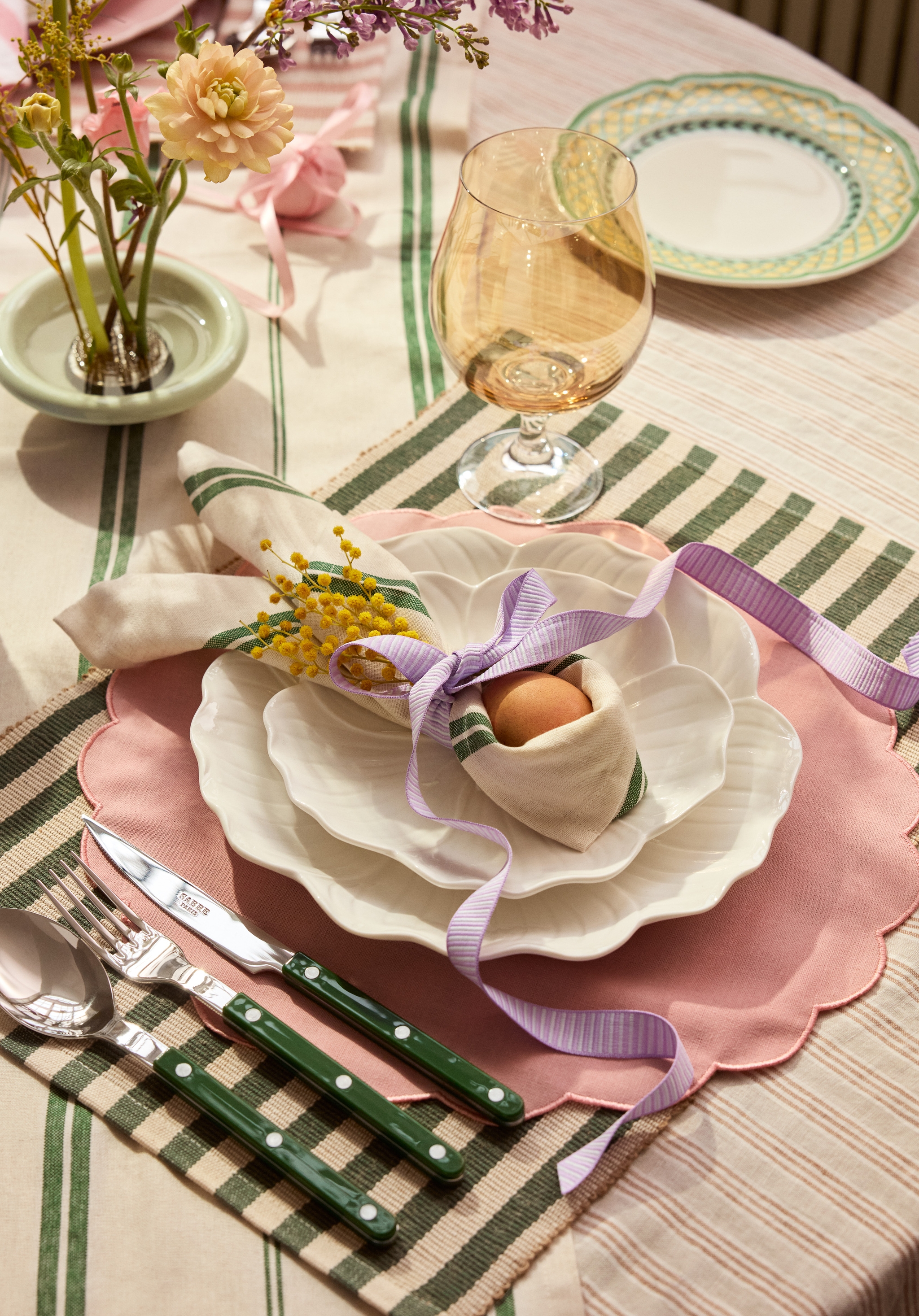 Keväinen kattaus, jossa on valkoiset aaltoreunaiset lautaset vaaleanpunaisella pöytätabletilla, raidallinen servetti, jossa on keltaista mimosa-kukkaa ja muna sidottuna violetilla nauhalla, sekä vihreävartiset aterimet