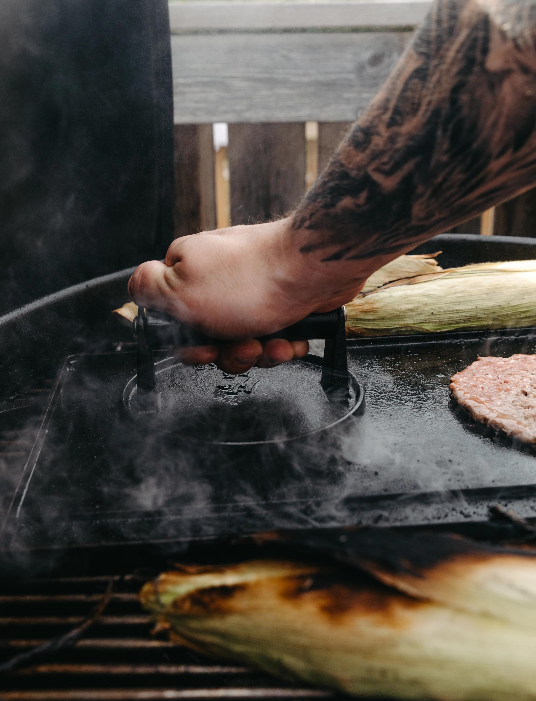 En tatuerad arm pressar en grillpress mot en hamburgare på en rykande grill bredvid majskolvar.