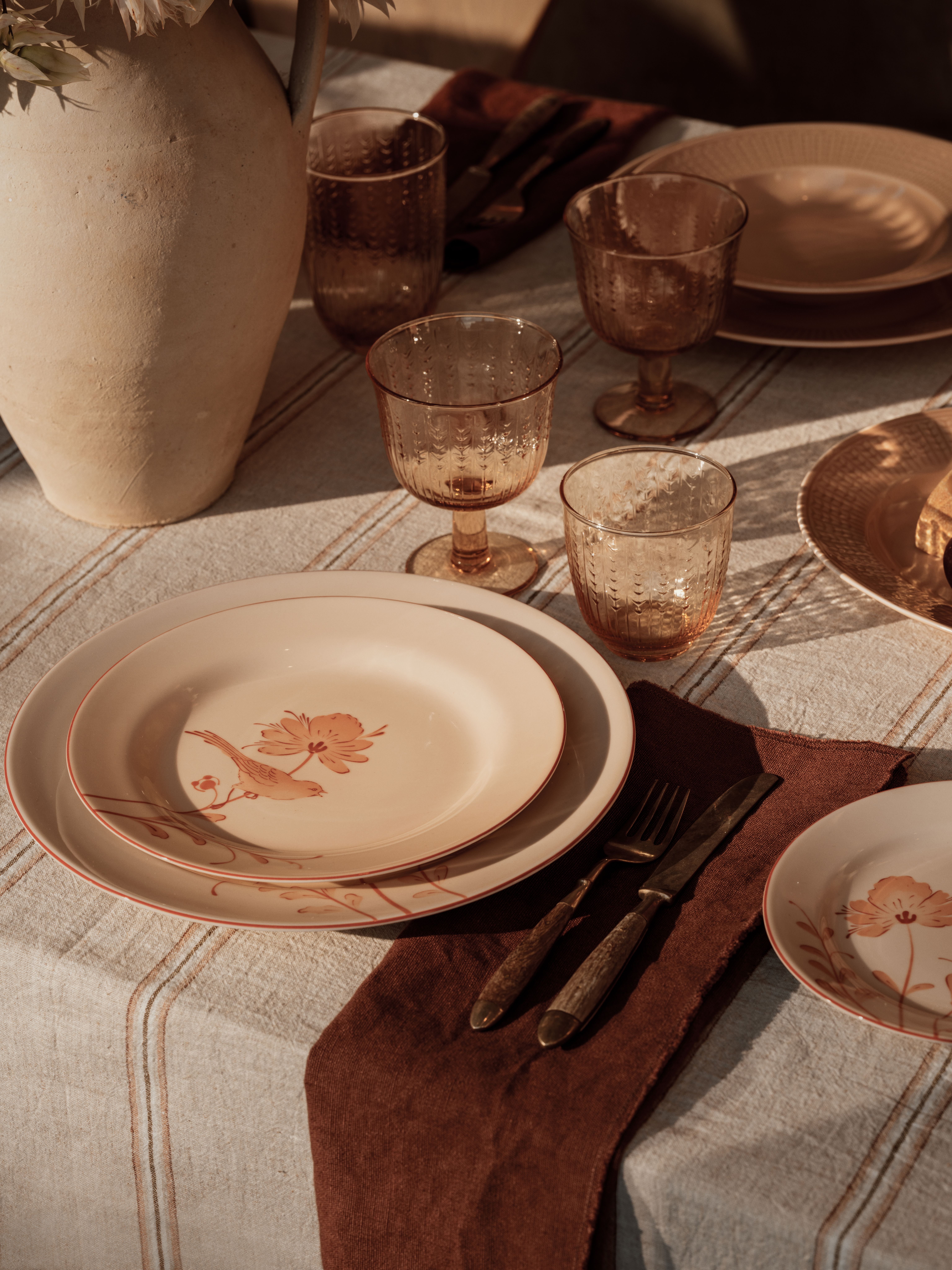 En borddekking i vintagestil med tallerkener med fugler og blomster, teksturerte ravfargede glass og antikt bestikk, opplyst av varmt sollys