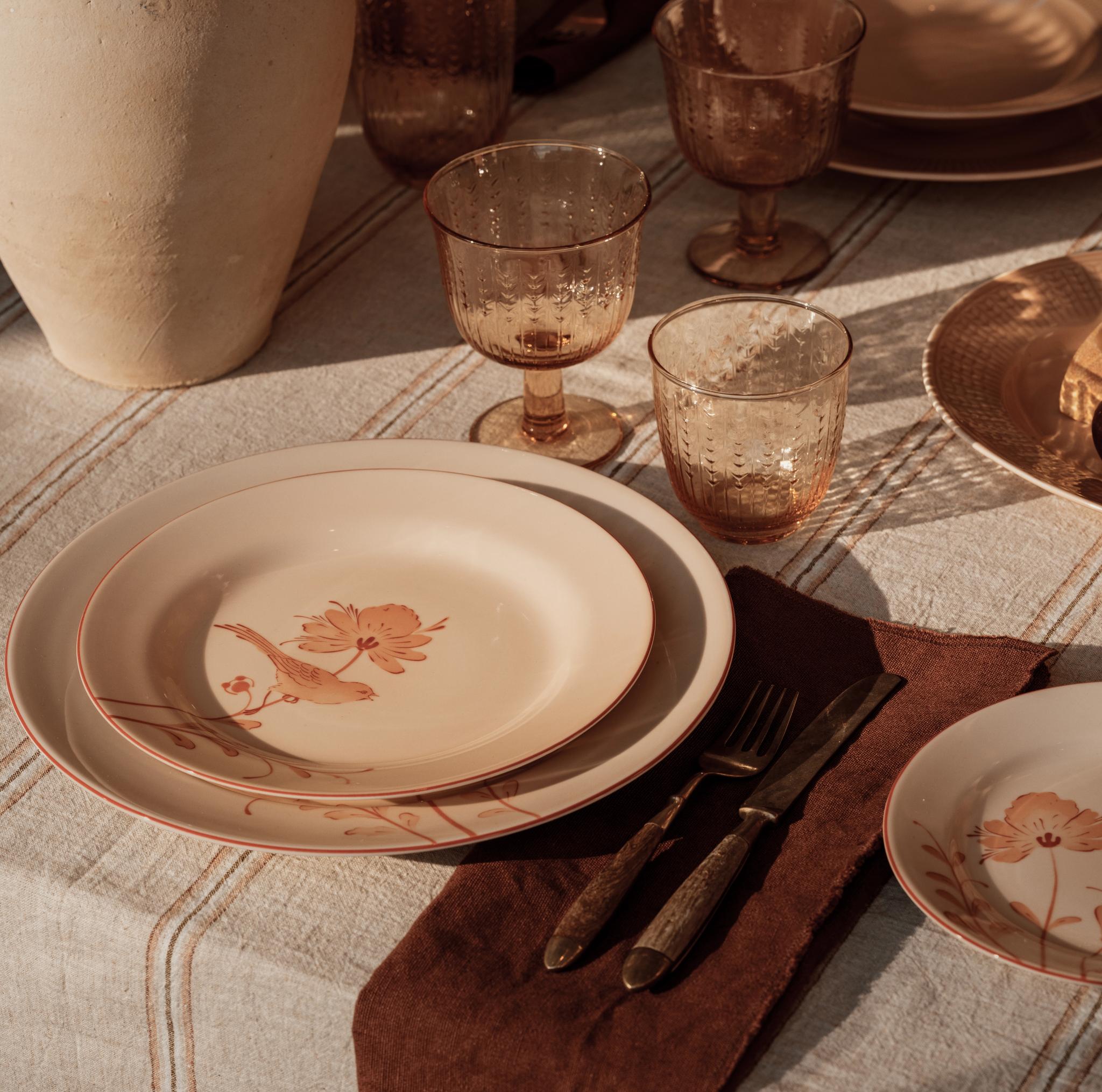 En borddekking i vintagestil med tallerkener med fugler og blomster, teksturerte ravfargede glass og antikt bestikk, opplyst av varmt sollys