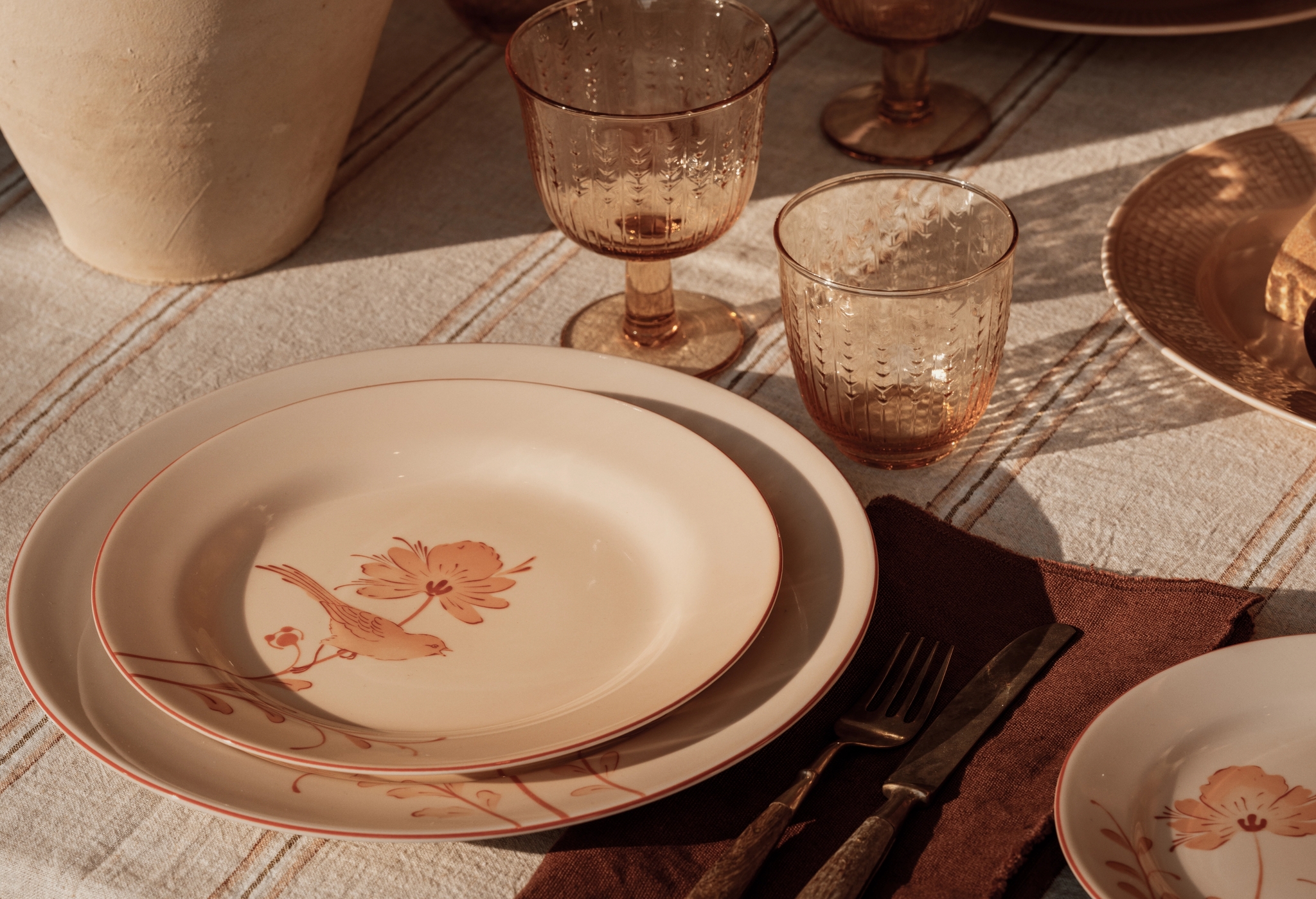 En borddekking i vintagestil med tallerkener med fugler og blomster, teksturerte ravfargede glass og antikt bestikk, opplyst av varmt sollys