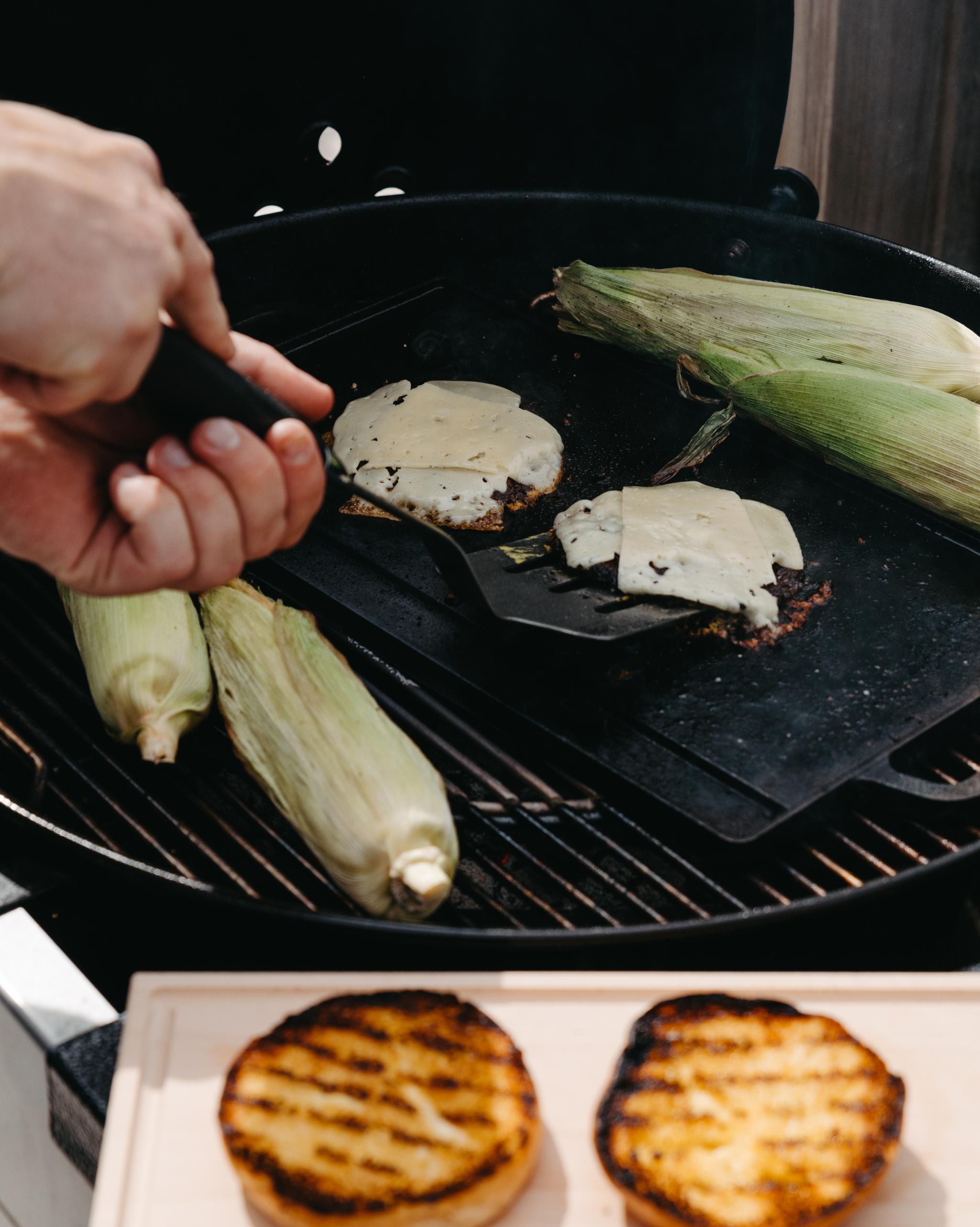 Hand som grillar hamburgare med smält ost, majskolvar och rostade hamburgerbröd på en kolgrill.