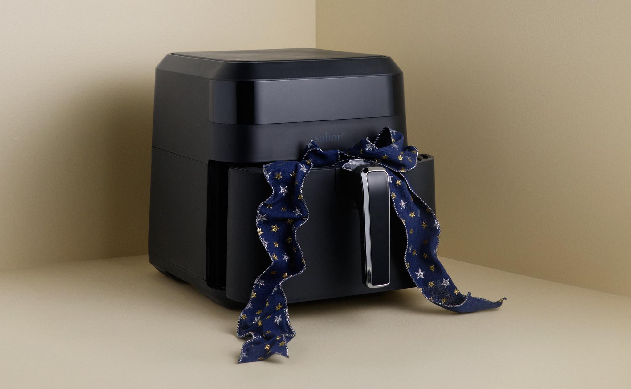 En svart airfryer med ett blått band knutet runt handtaget, placerad i ett ljusbeige hörn.