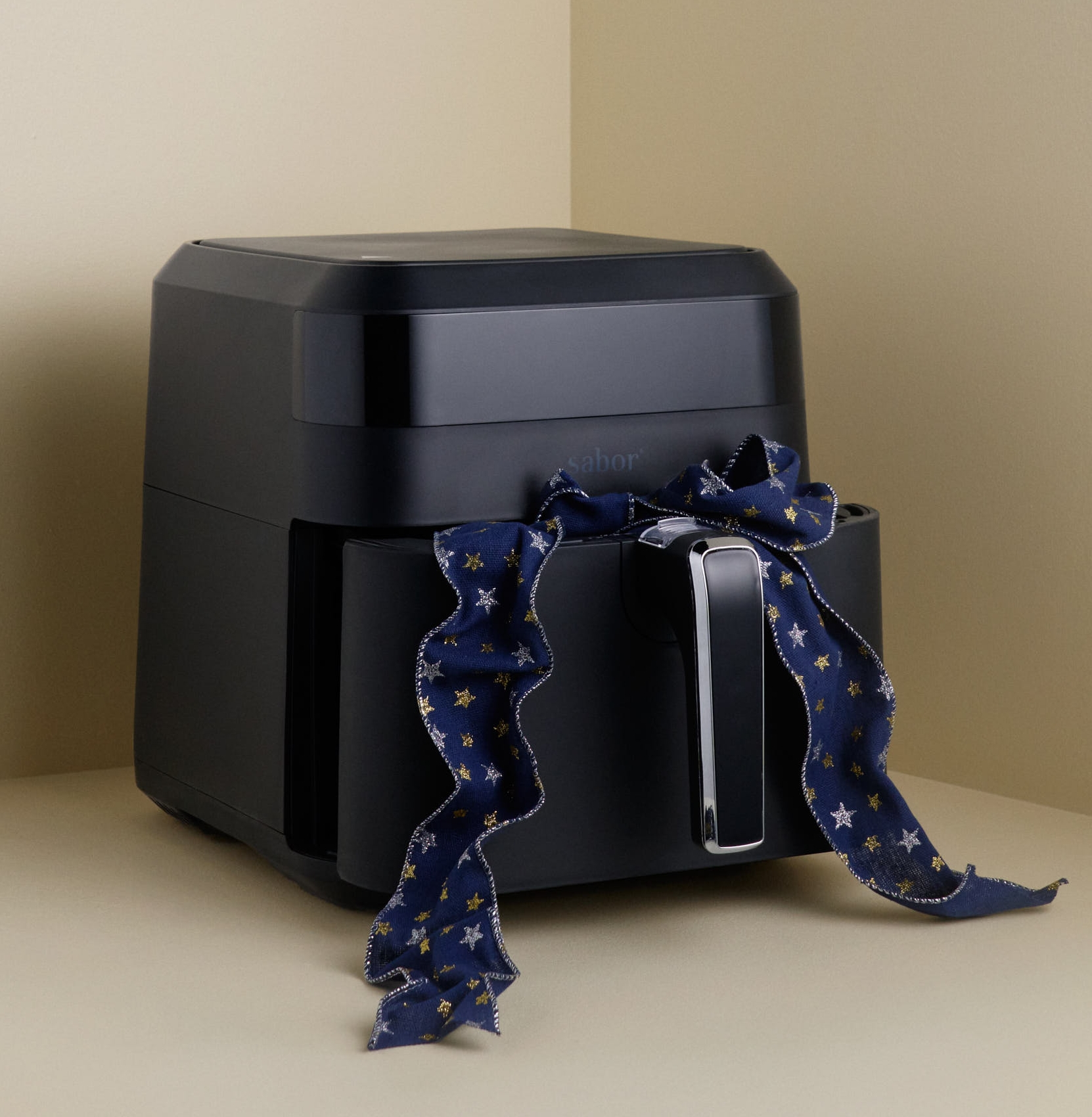 En svart airfryer med ett blått band knutet runt handtaget, placerad i ett ljusbeige hörn.