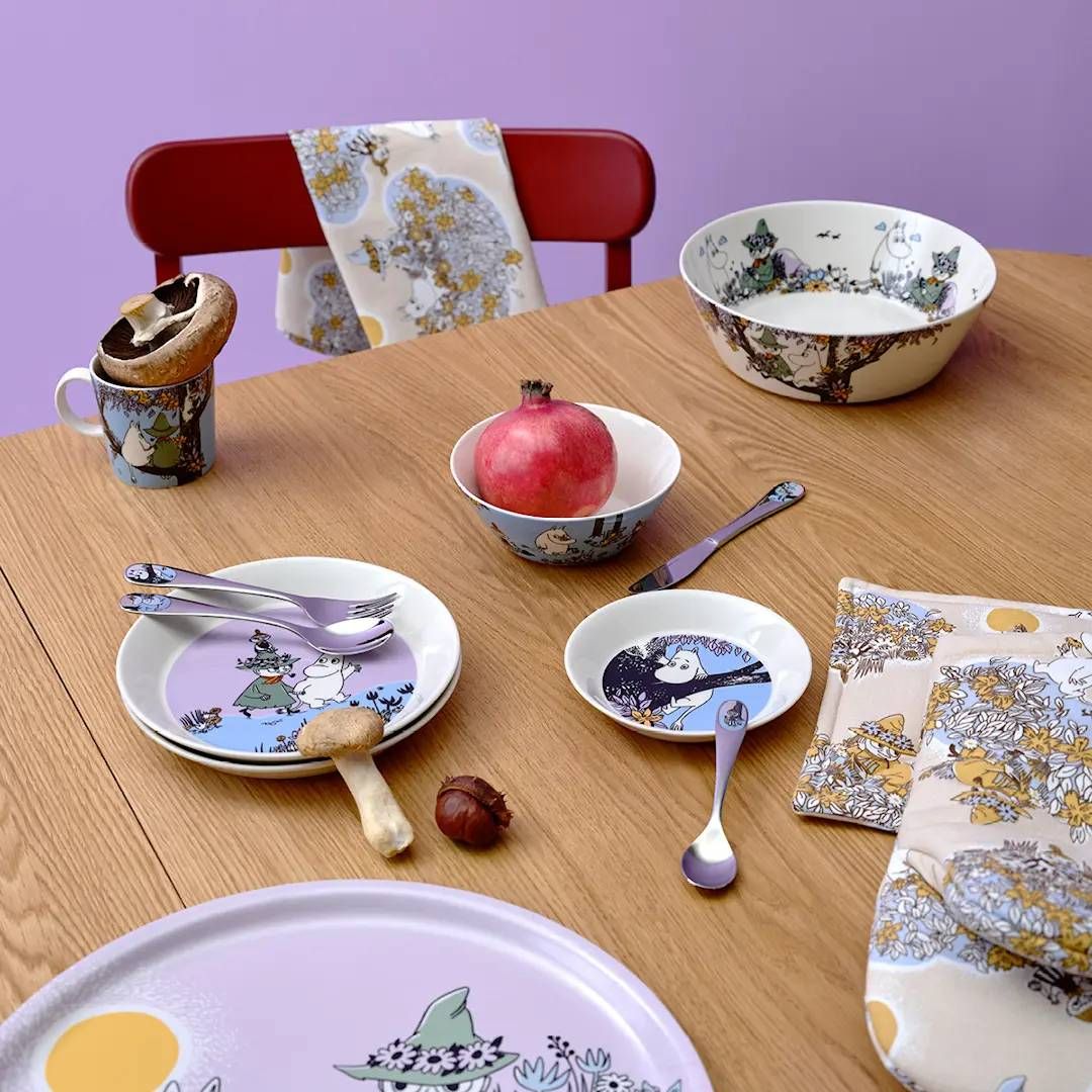 Muumi-astiasto, jossa on lautasia, kulhoja, mukeja ja aterimia puisella pöydällä granaattiomenan ja sienten kanssa violetilla taustalla