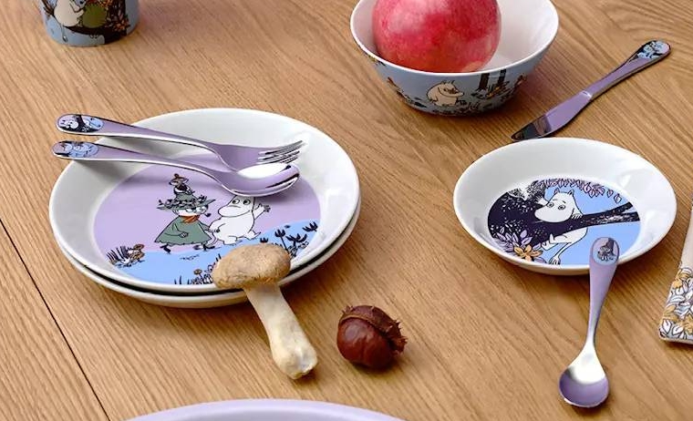 Muumi-astiasto, jossa on lautasia, kulhoja, mukeja ja aterimia puisella pöydällä granaattiomenan ja sienten kanssa violetilla taustalla