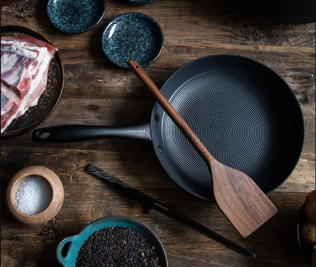 En mörk stekpanna med mönstrad yta och en träspatel inuti, på ett rustikt träbord.
