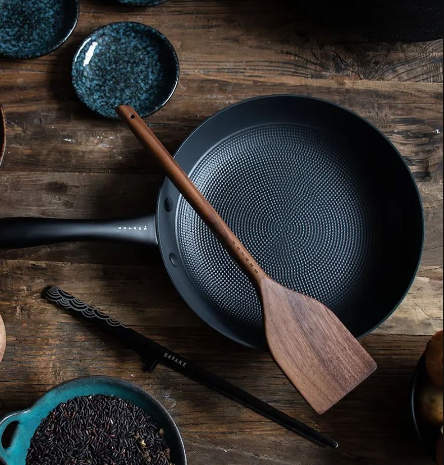 En mörk stekpanna med mönstrad yta och en träspatel inuti, på ett rustikt träbord.