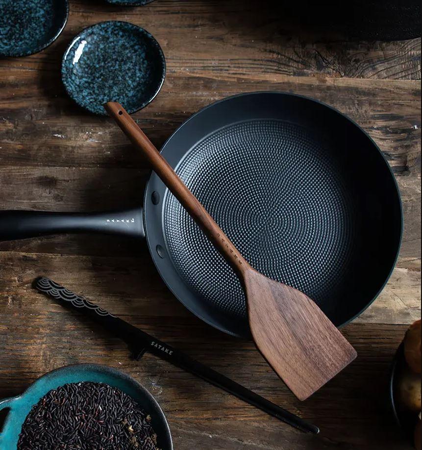 En mörk stekpanna med mönstrad yta och en träspatel inuti, på ett rustikt träbord.