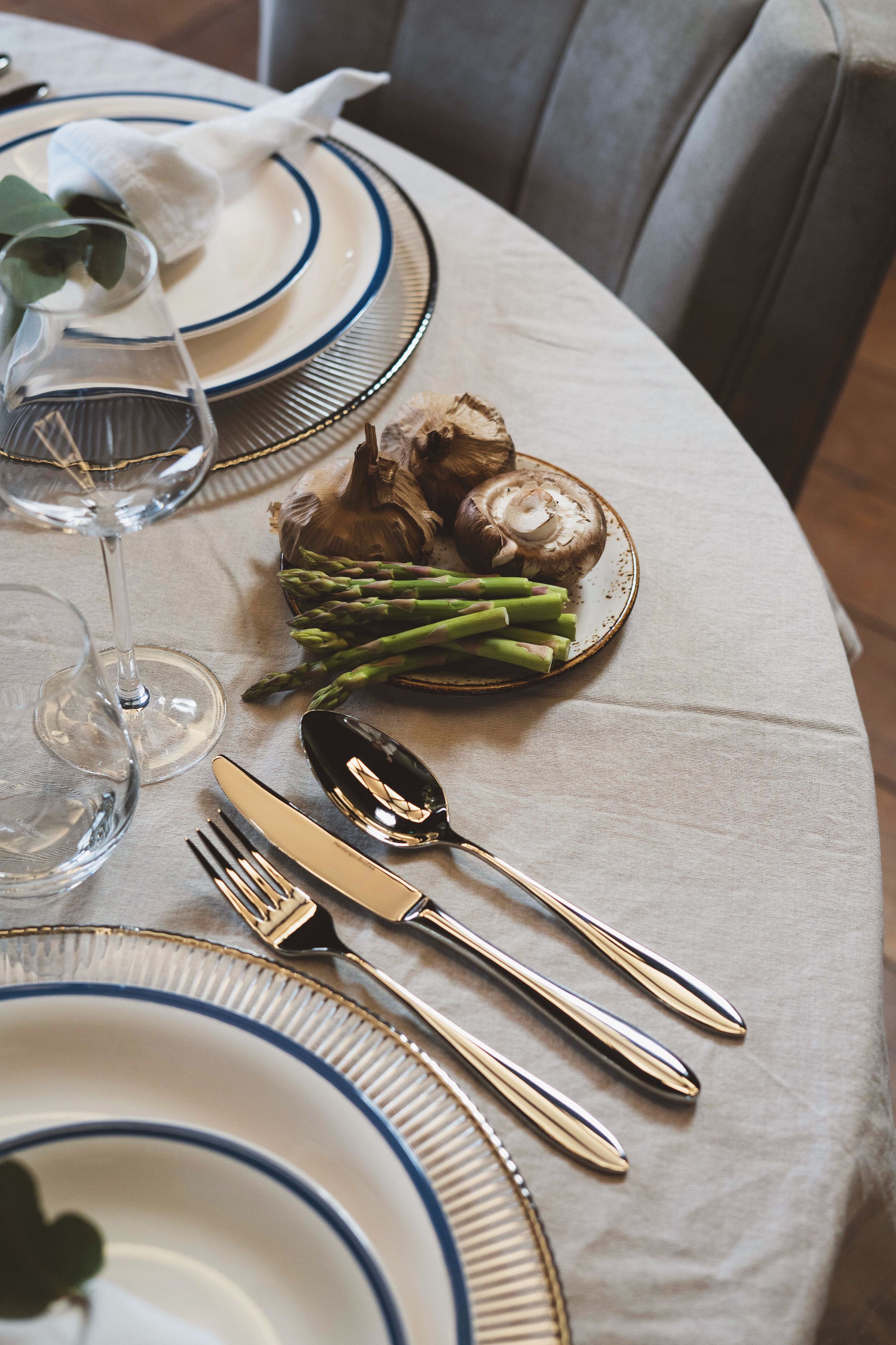 Ett matbord dukat med tallrikar med blå kant, bestick, glas och en fat med färsk sparris, svamp och vitlök.