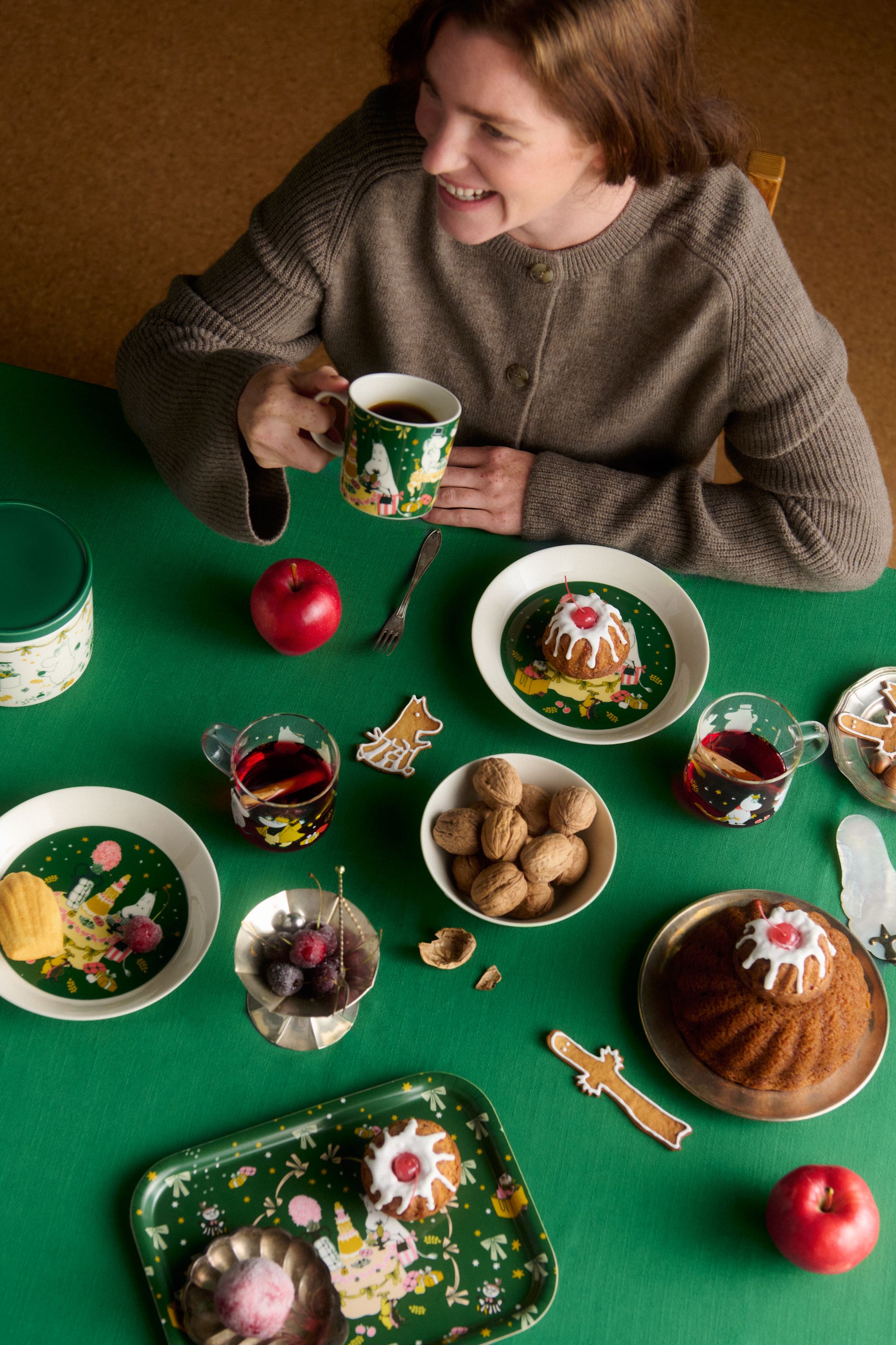 Leende kvinna njuter av kaffe vid ett juldukat bord med bakverk, pepparkakor, äpplen och Muminservis.