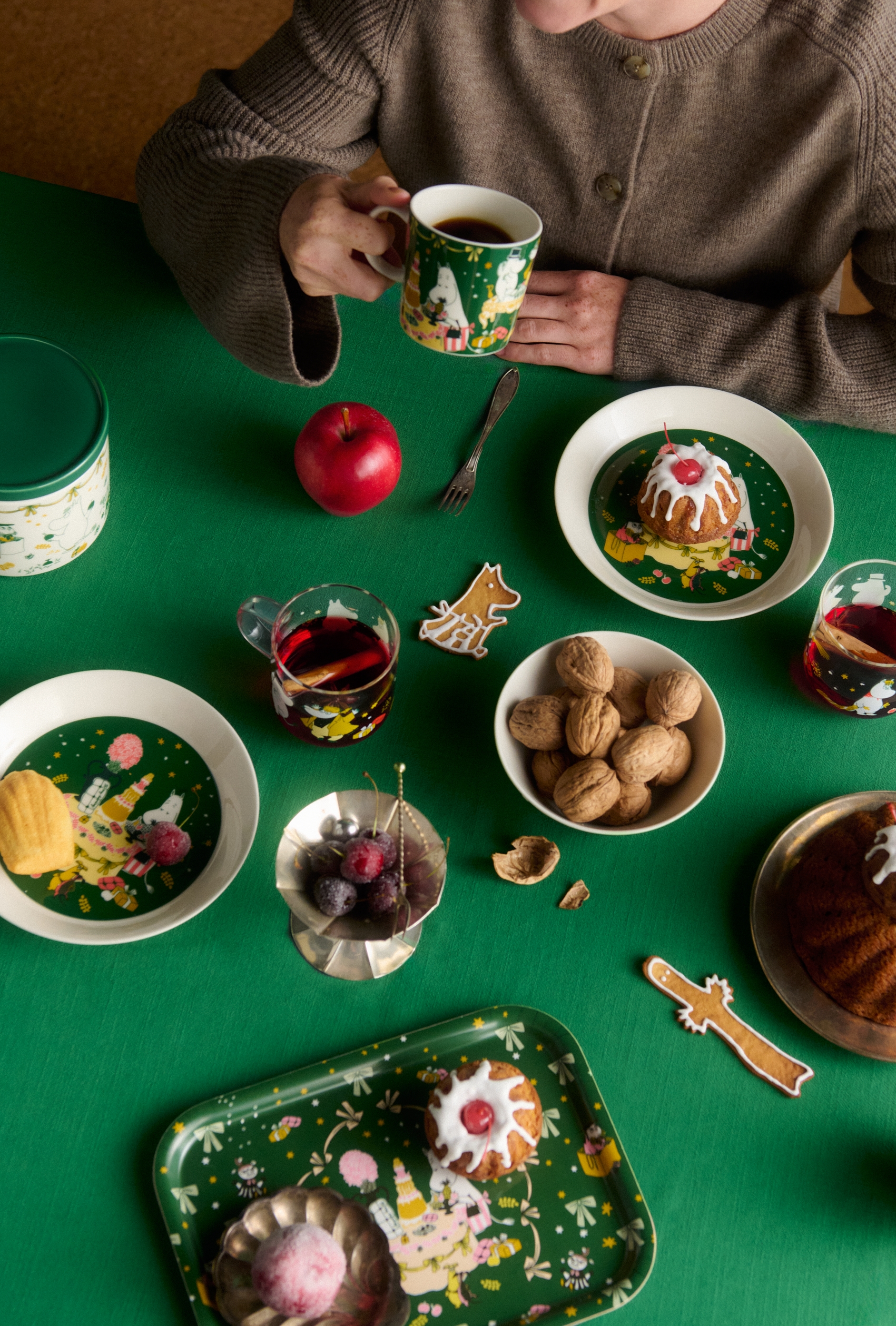 Leende kvinna njuter av kaffe vid ett juldukat bord med bakverk, pepparkakor, äpplen och Muminservis.