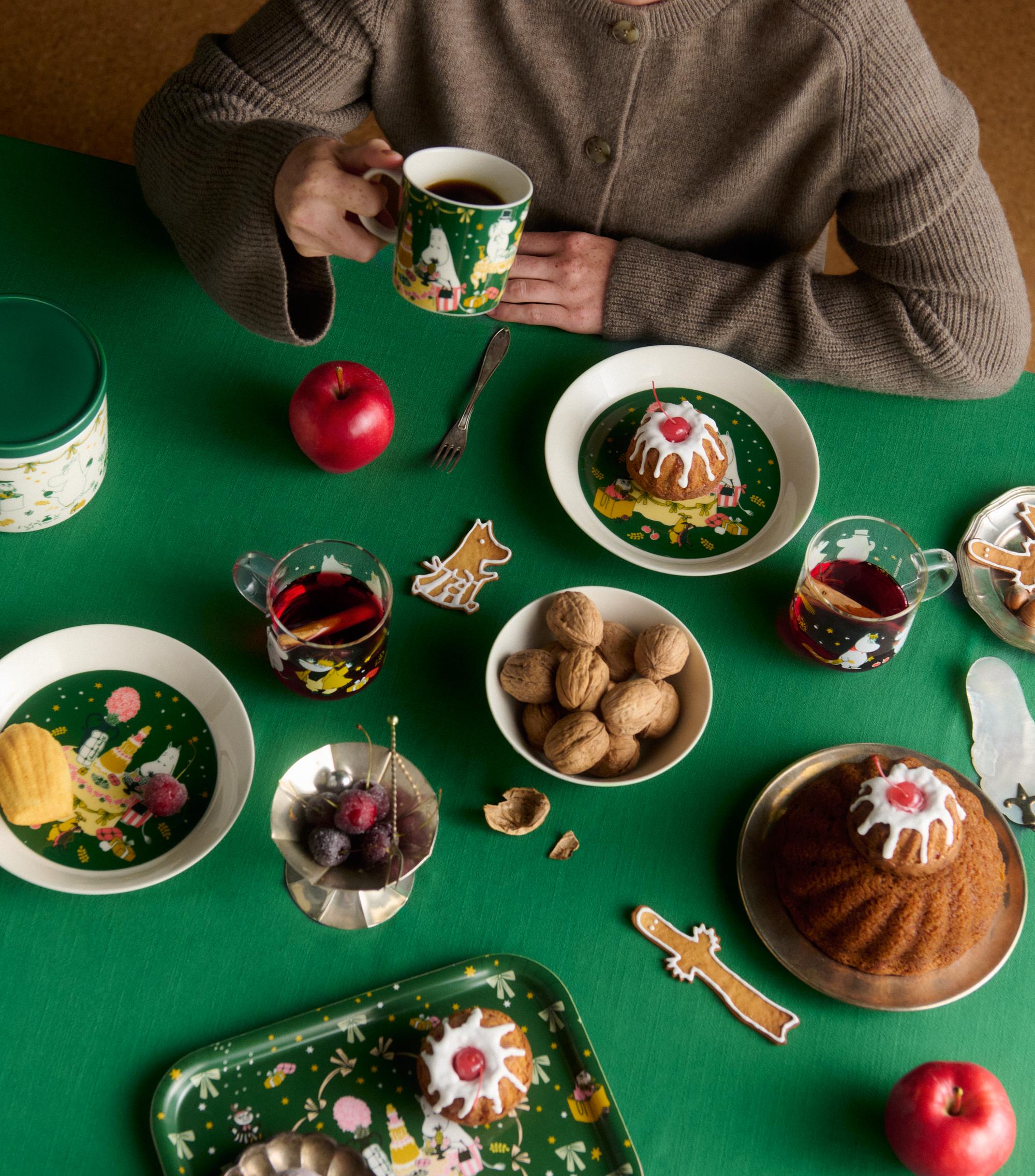 Leende kvinna njuter av kaffe vid ett juldukat bord med bakverk, pepparkakor, äpplen och Muminservis.