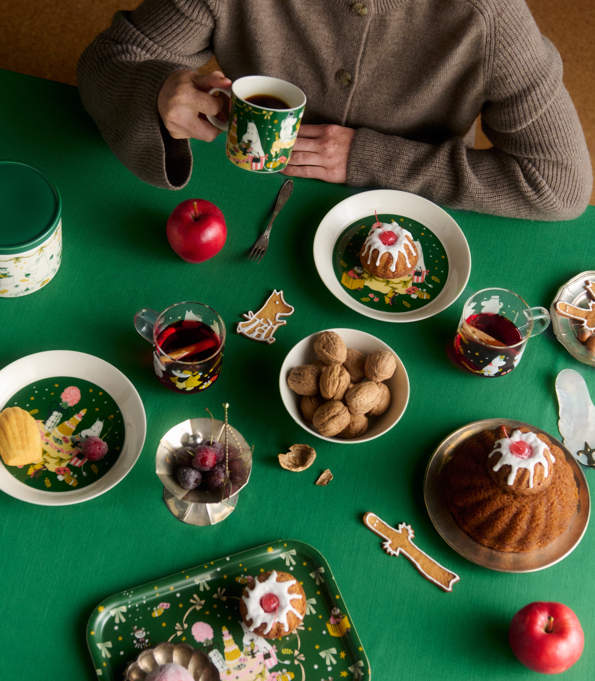 Leende kvinna med kaffekopp vid ett juldukat bord med kakor, glögg, äpplen och nötter.