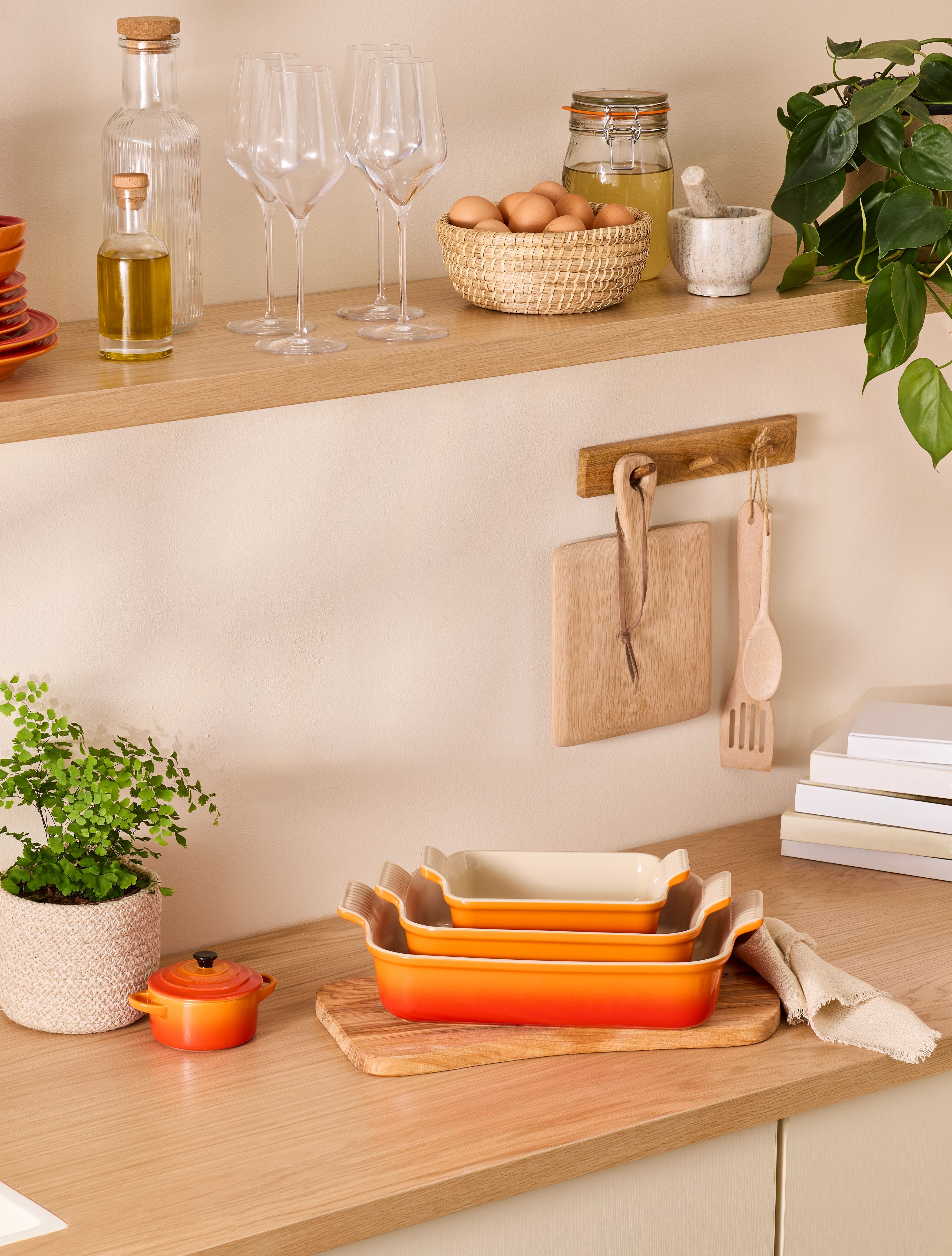 Stilren köksbänk med staplade orange ugnsformar, örter, äggkorg och glas på en trähylla i ett ljust, modernt kök.