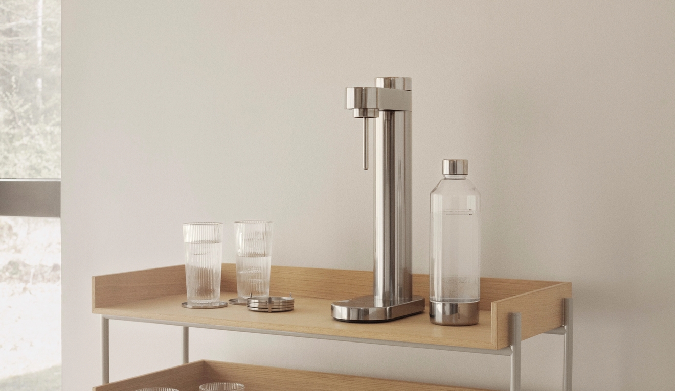 En modern kolsyremaskin i rostfritt stål, en glasflaska och glas på en ljus trähylla.