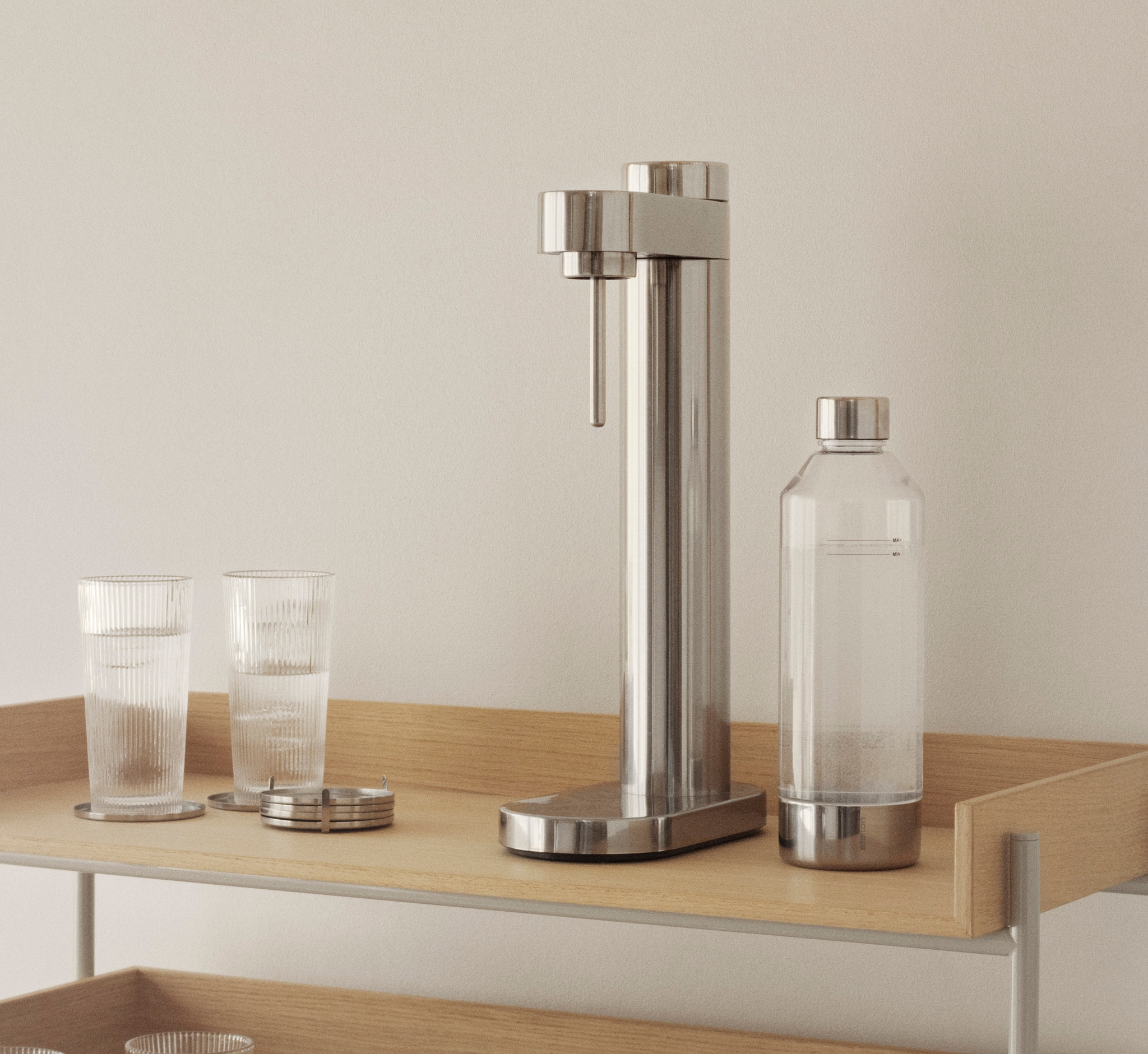 En modern trähylla i tre våningar med en silverfärgad kolsyremaskin, glas, böcker och trämuggar.