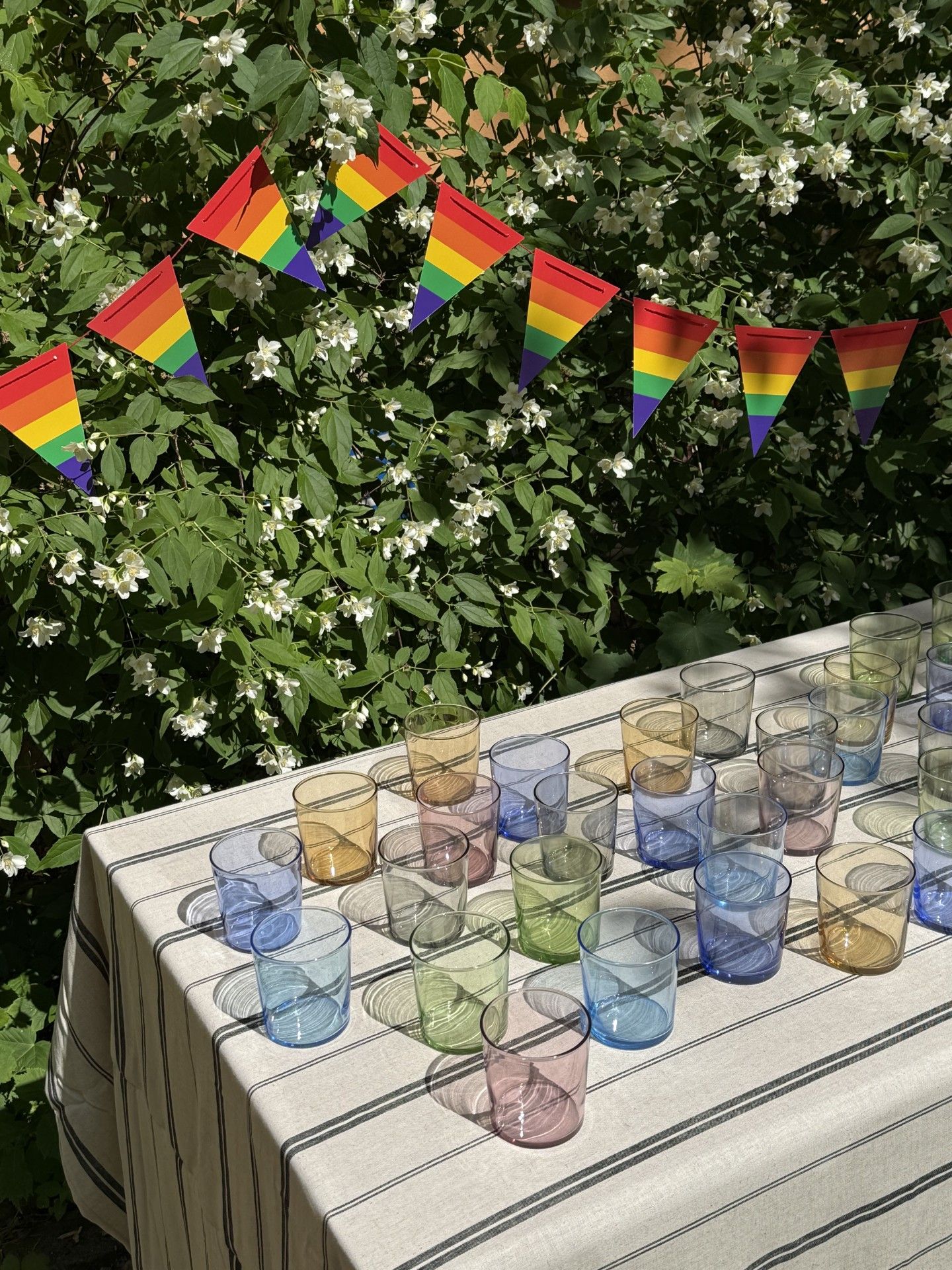 Färgglada glas står på ett dukat bord utomhus framför gröna buskar med vita blommor och en girlang med regnbågsflaggor.