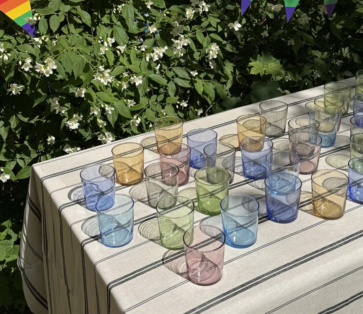 Färgglada glas står på ett dukat bord utomhus framför gröna buskar med vita blommor och en girlang med regnbågsflaggor.