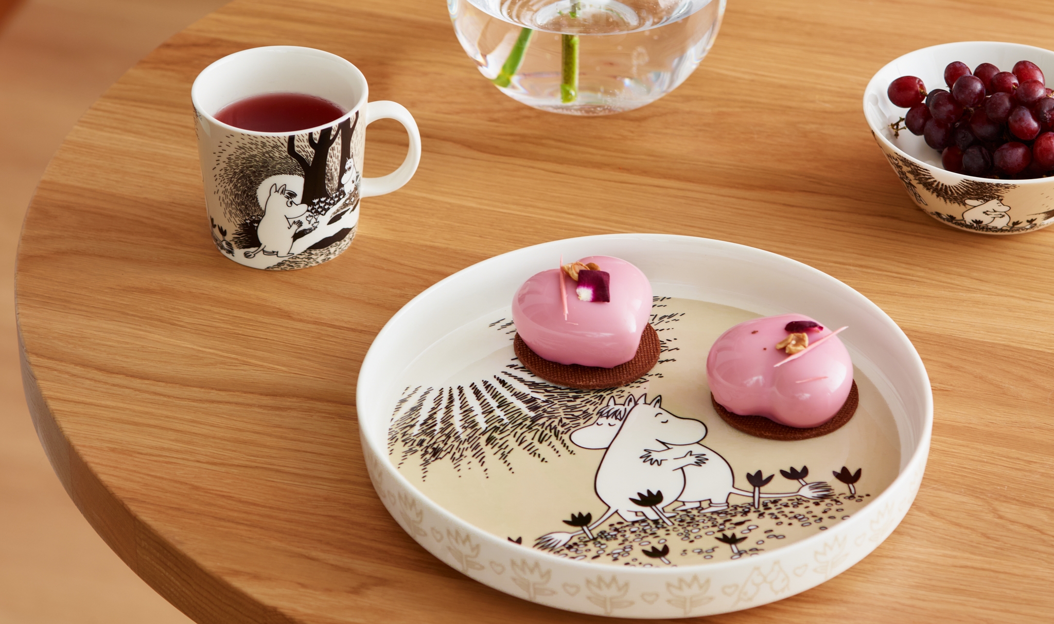 Dukat träbord med Mumin-porslin, rosa bakelser, en kopp te och en glasvas med blomma i ett stilrent hem.