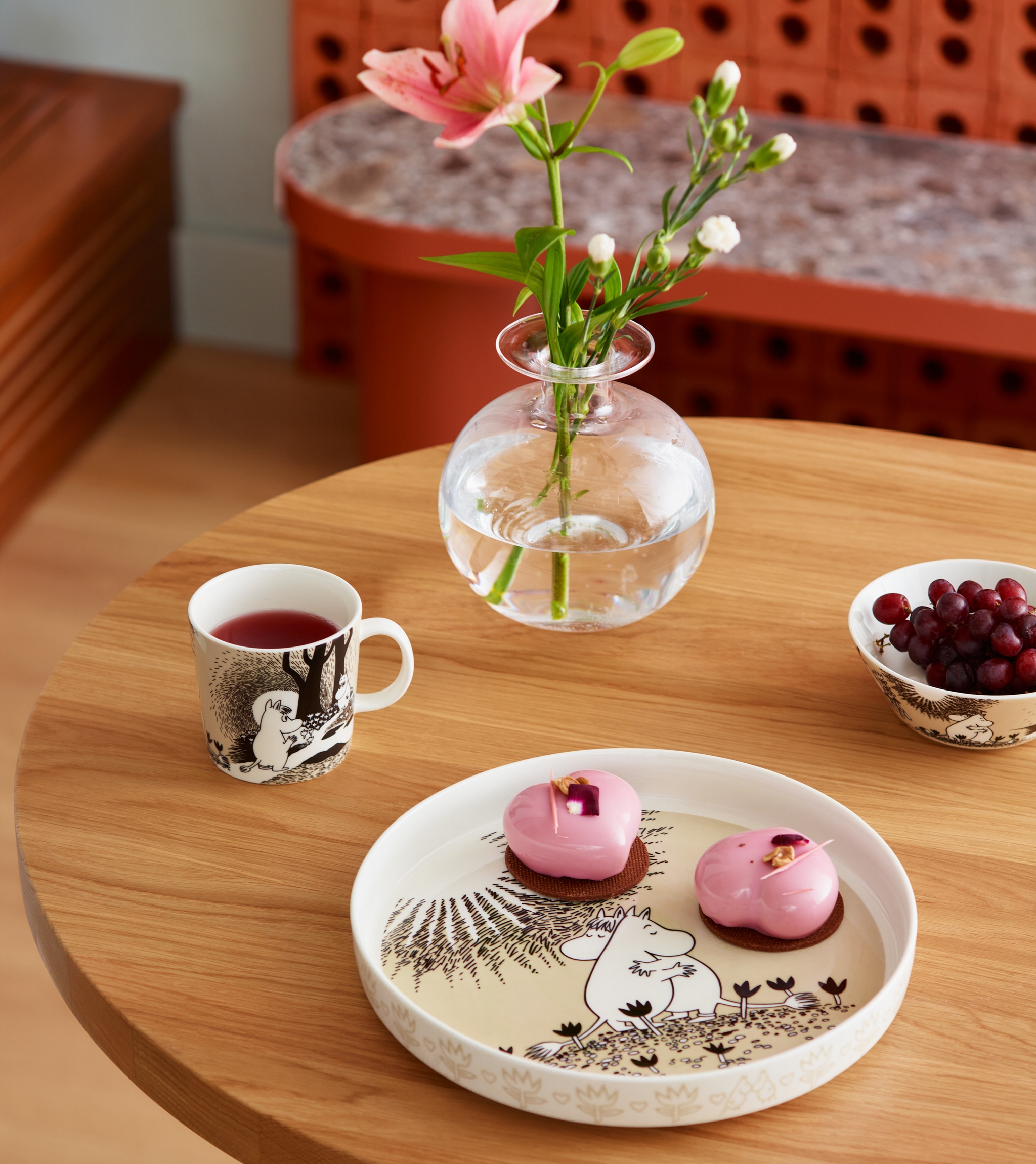 Dukat träbord med Mumin-porslin, rosa bakelser, en kopp te och en glasvas med blomma i ett stilrent kök.