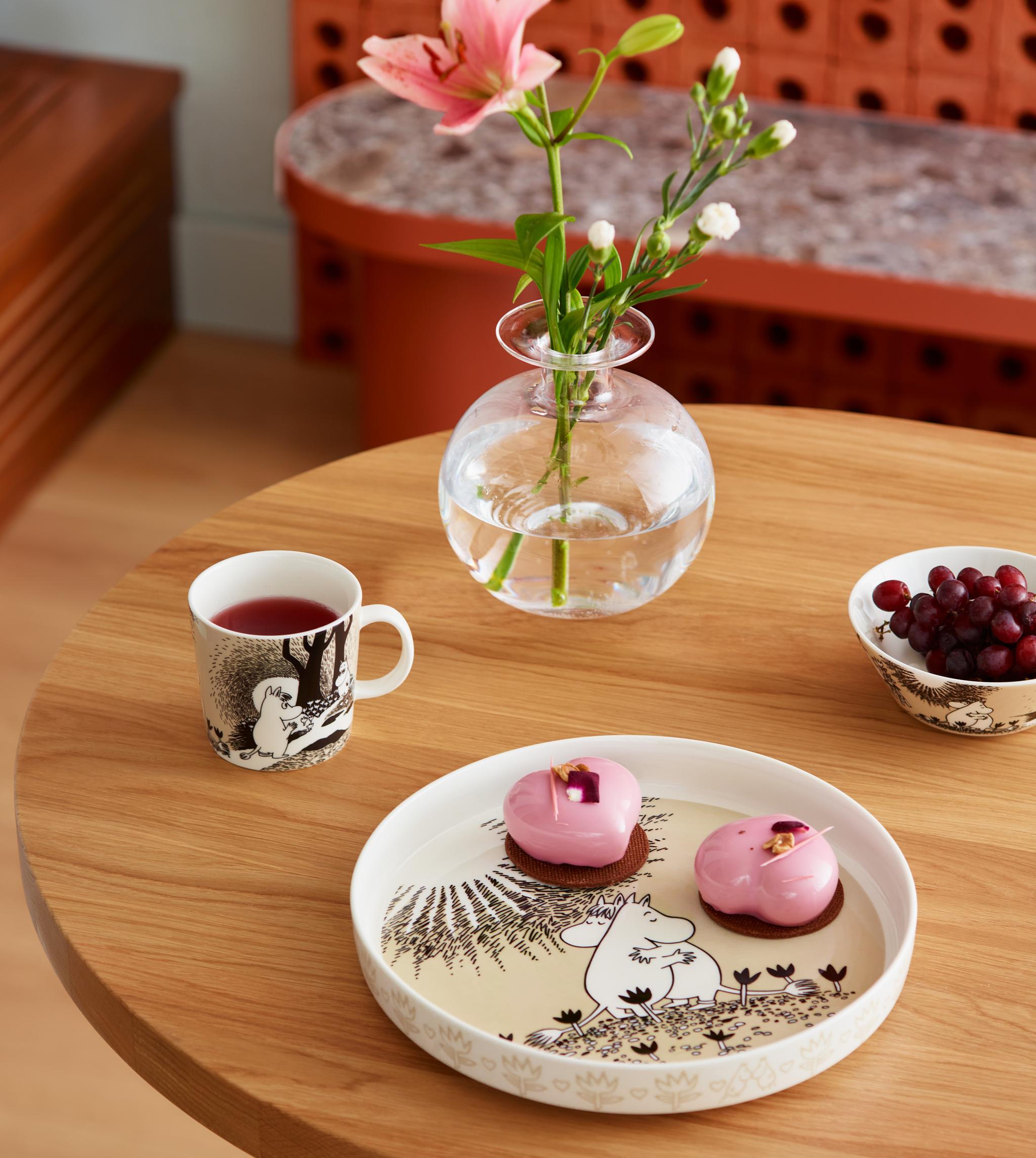 Dukat träbord med Mumin-porslin, rosa bakelser, en kopp te och en glasvas med blomma skapar en mysig fikastund.