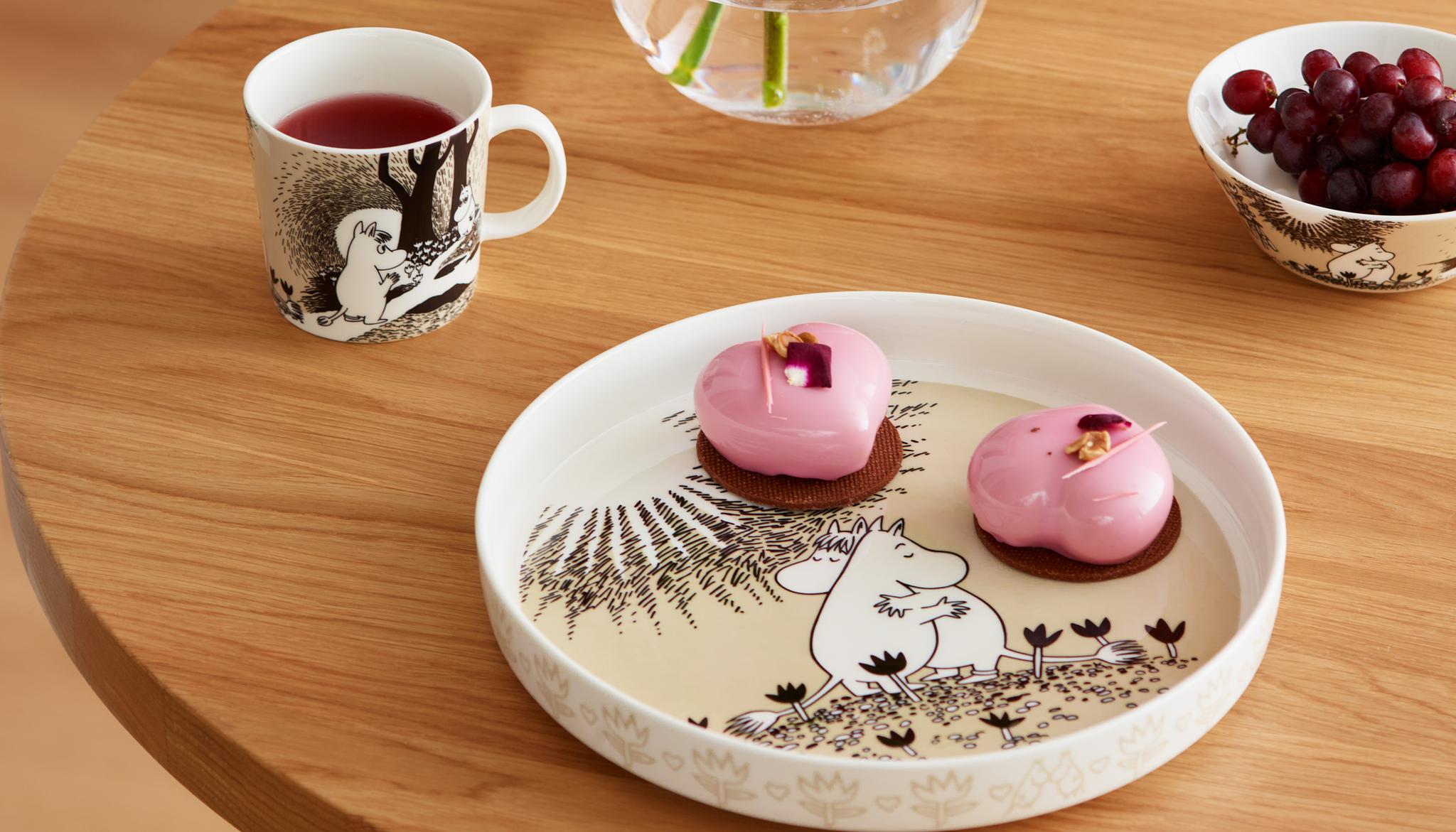 Runt träbord dukat med Mumin-porslin, rosa bakelser, en kopp te och en glasvas med blomma i ett stilrent hem.