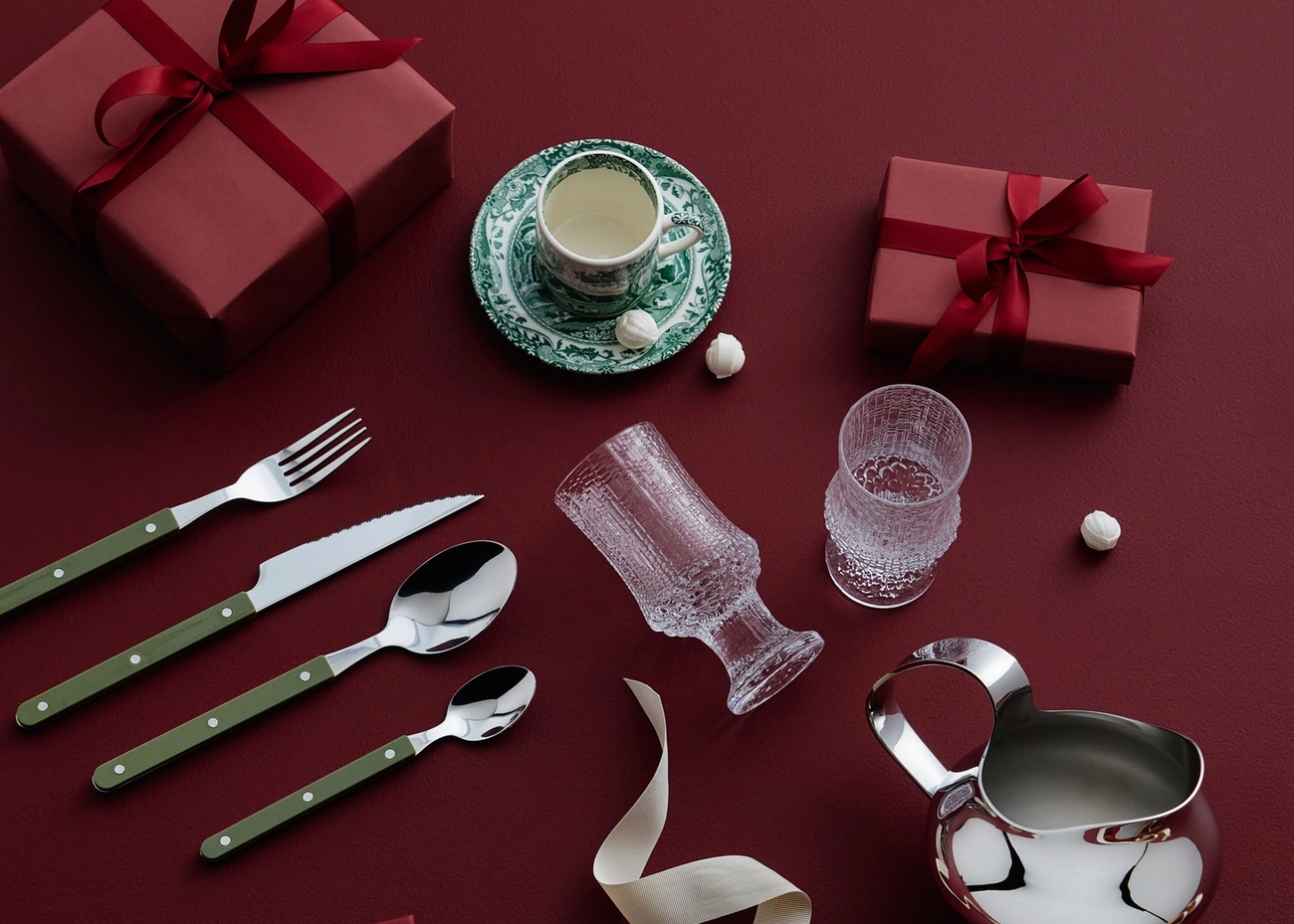 Röda presentaskar, bestick med gröna handtag, en tekopp och glas arrangerade på en mörkröd yta.