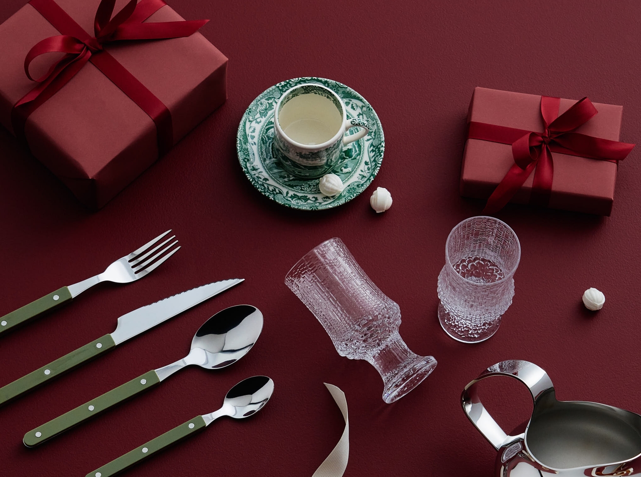 Röda presentaskar, bestick med gröna handtag, en tekopp och glas arrangerade på en mörkröd yta.