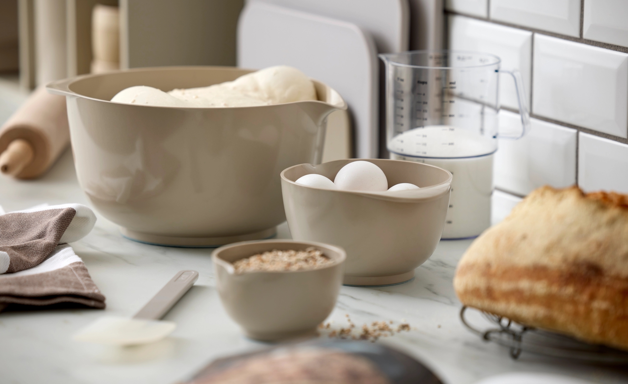 Bakeingredienser med brøddeig, egg, melk og mel står på en kjøkkenbenk ved siden av et nystekt brød.