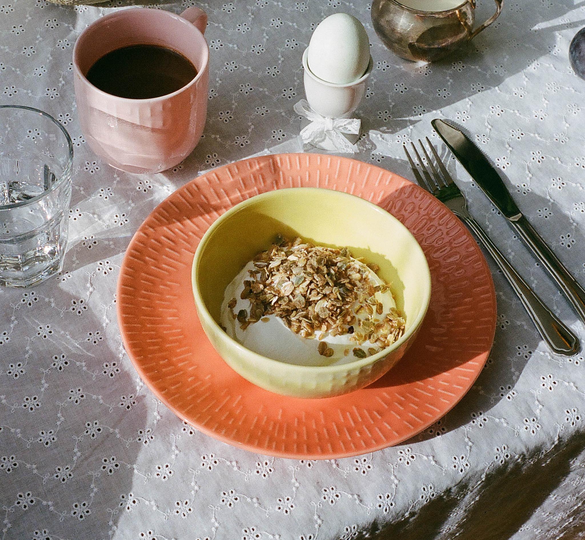 Vackert uppdukad frukost med yoghurt och müsli, kaffe, kokt ägg, melon med röda vinbär och ost på ett solbelyst bord.