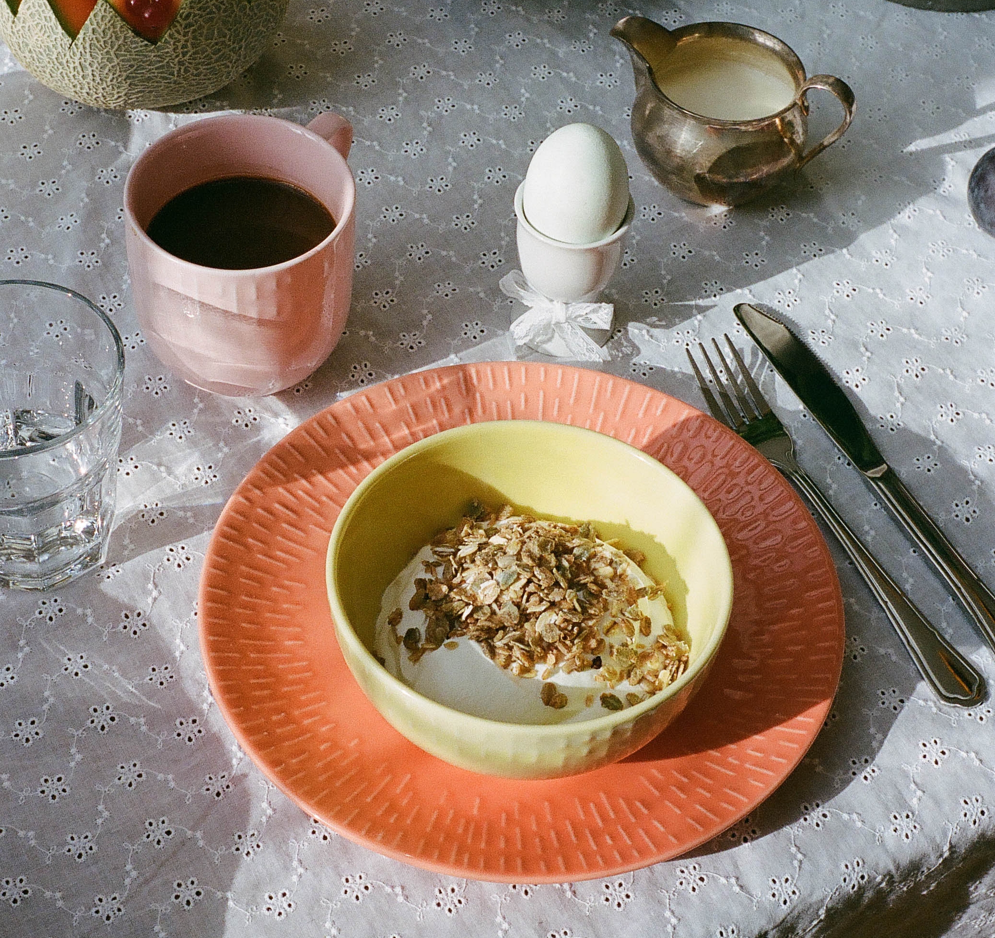 Somrig frukostdukning med yoghurt och müsli, kaffe, kokt ägg, melon med röda vinbär och ost på ett ljust spetsdukat bord.