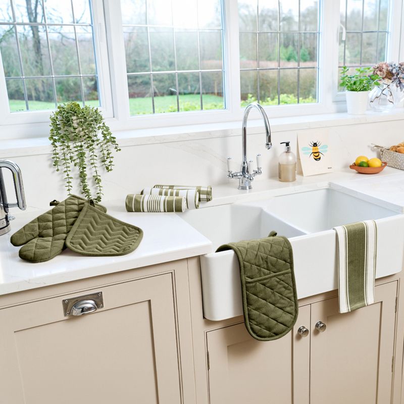 Vaaleassa keittiössä on suuri valkoinen allas, vihreät patalaput ja raidalliset keittiöpyyhkeet ikkunan alla.]}