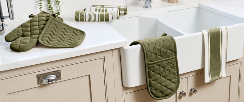 Vaaleassa keittiössä valkoisen altaan ympärillä on vihreitä patalappuja ja raidallisia keittiöpyyhkeitä ikkunan edessä avautuvaan puutarhanäkymään päin.