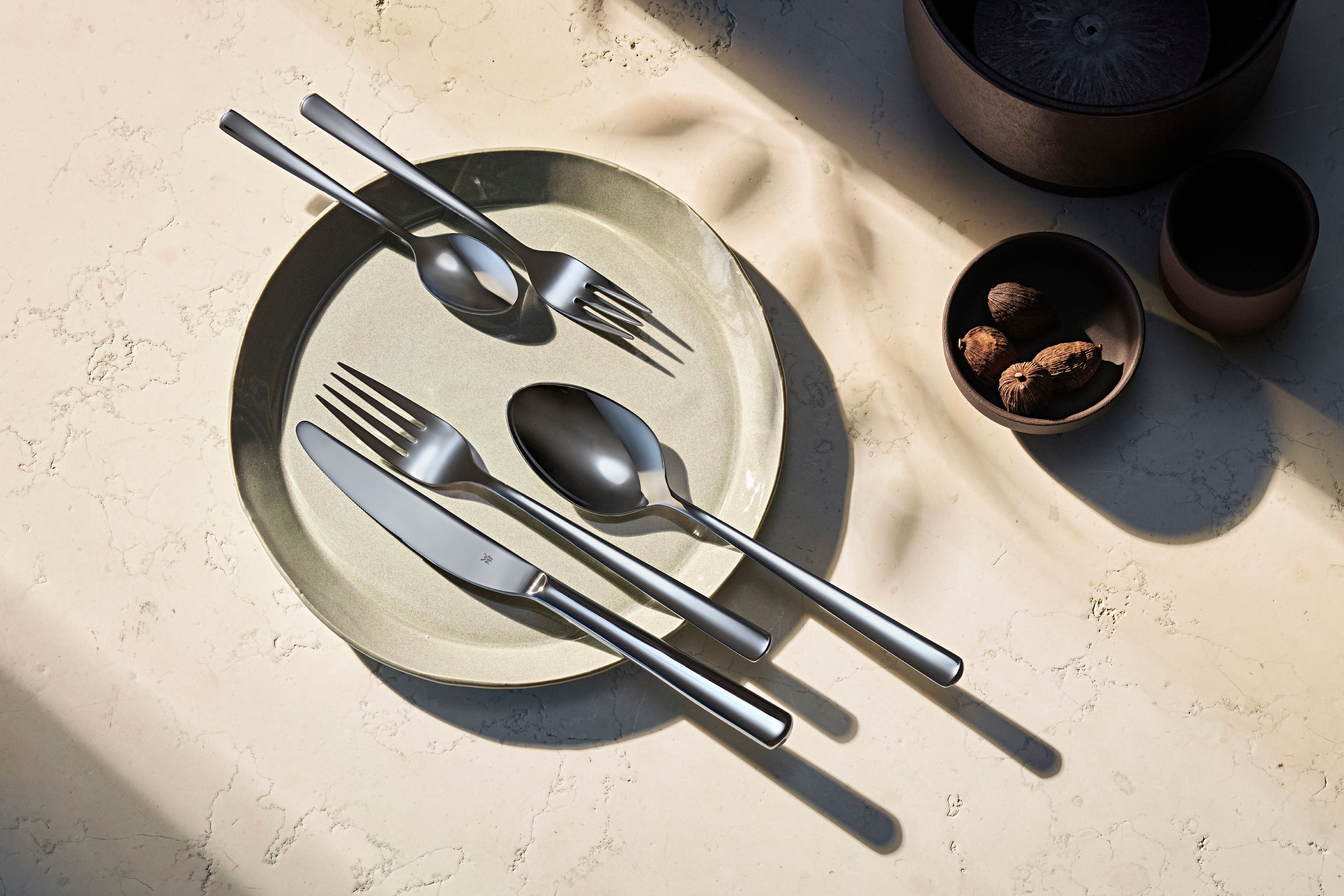 Moderneja aterimia vaaleanvihreällä lautasella, pieni kulho pähkinöitä ja voimakkaita varjoja.