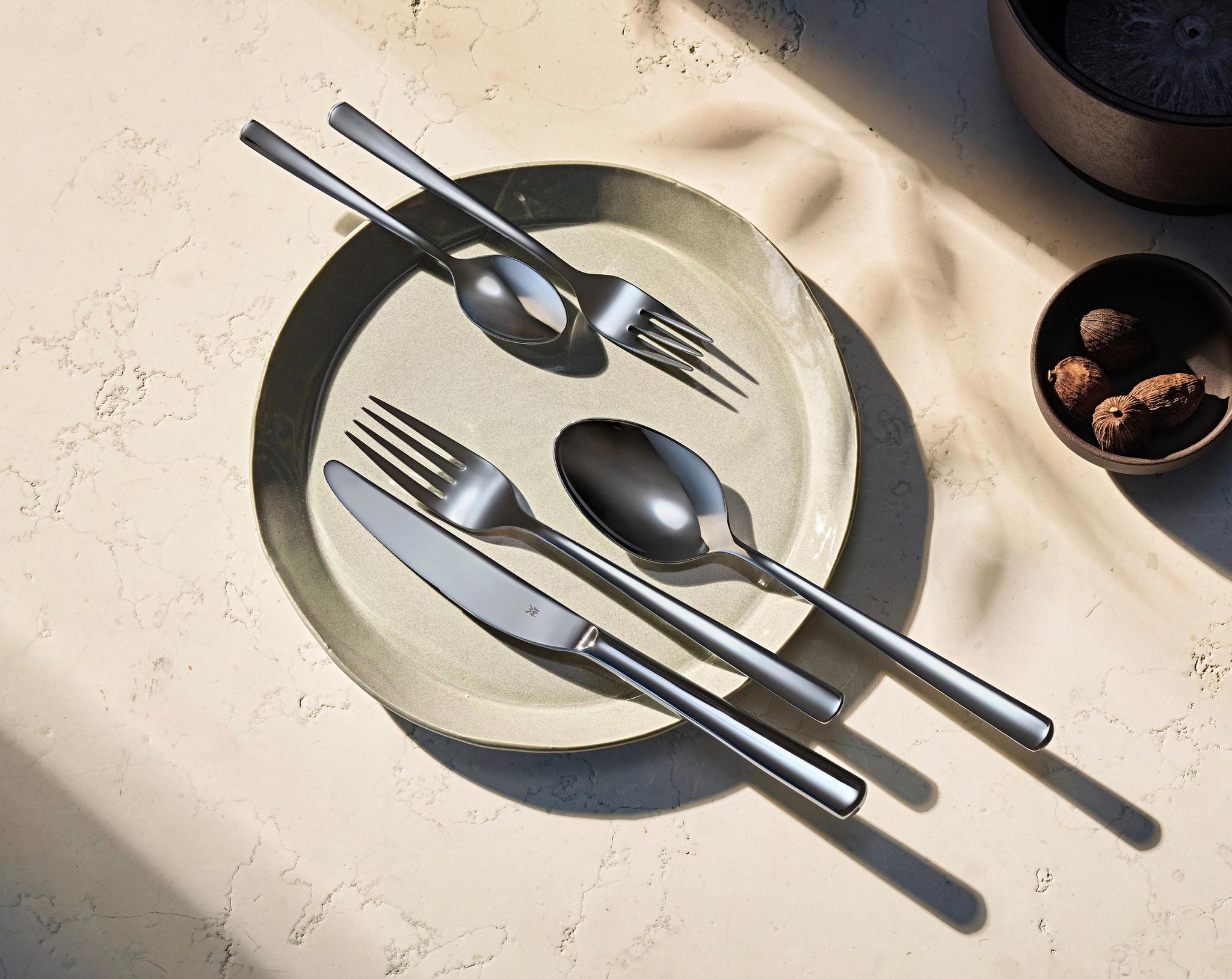 Moderneja aterimia vaaleanvihreällä lautasella, pieni kulho pähkinöitä ja voimakkaita varjoja.