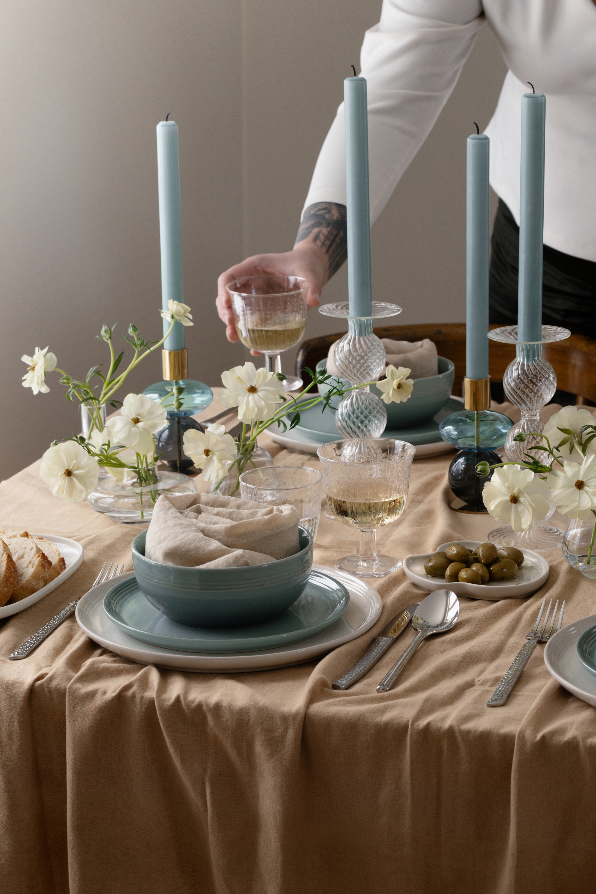En persons hand sträcker sig efter ett vinglas på ett elegant dukat bord med blå ljus, vita blommor och porslin.