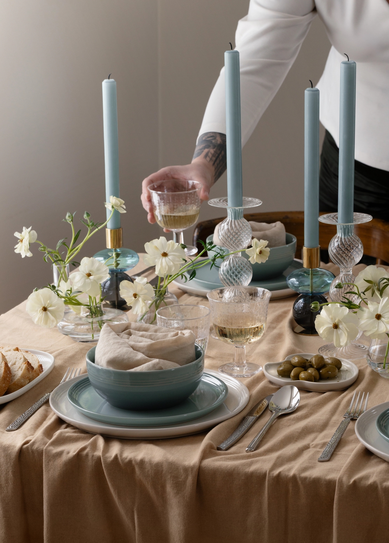 En persons arm sträcker sig efter ett vinglas på ett matbord dukat med blå ljus, vita blommor och blåvitt porslin ovanpå en beige duk.