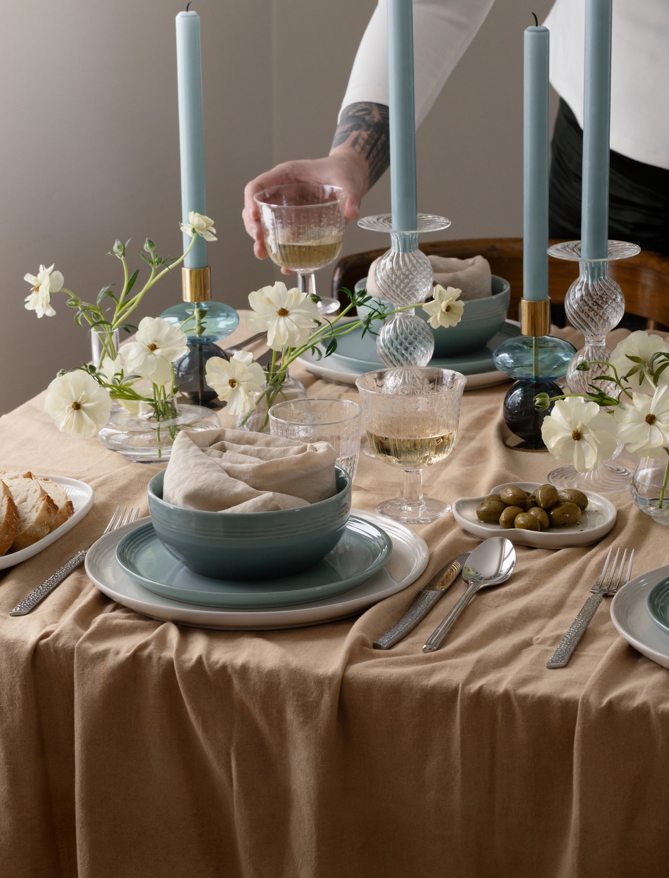 En persons arm sträcker sig efter ett vinglas på ett matbord dukat med blå ljus, vita blommor och blåvitt porslin ovanpå en beige duk.