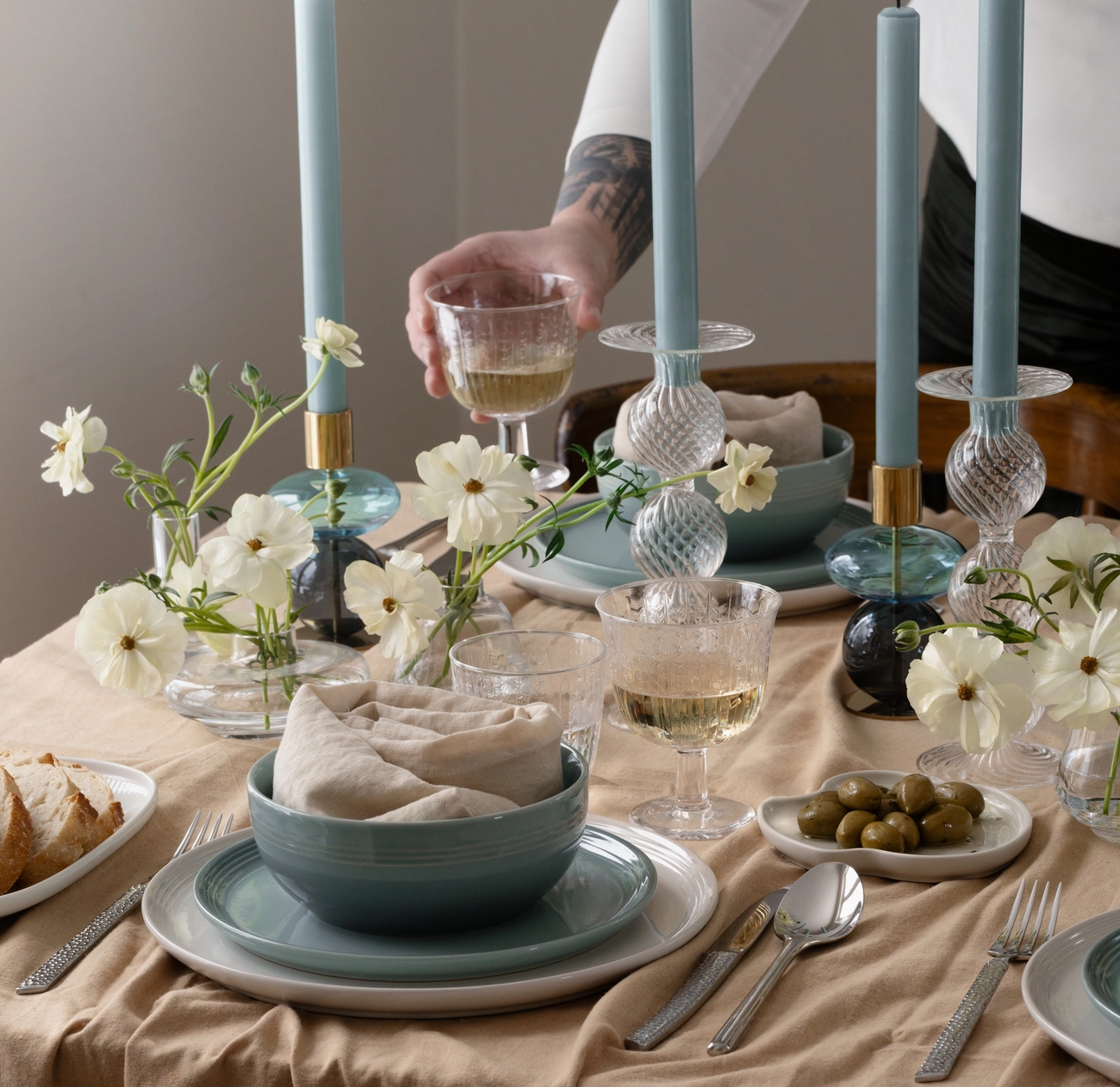 A set dining table with a person's hand holding a wine glass, tall blue candles, white flowers, blue dishes, bread, and olives.