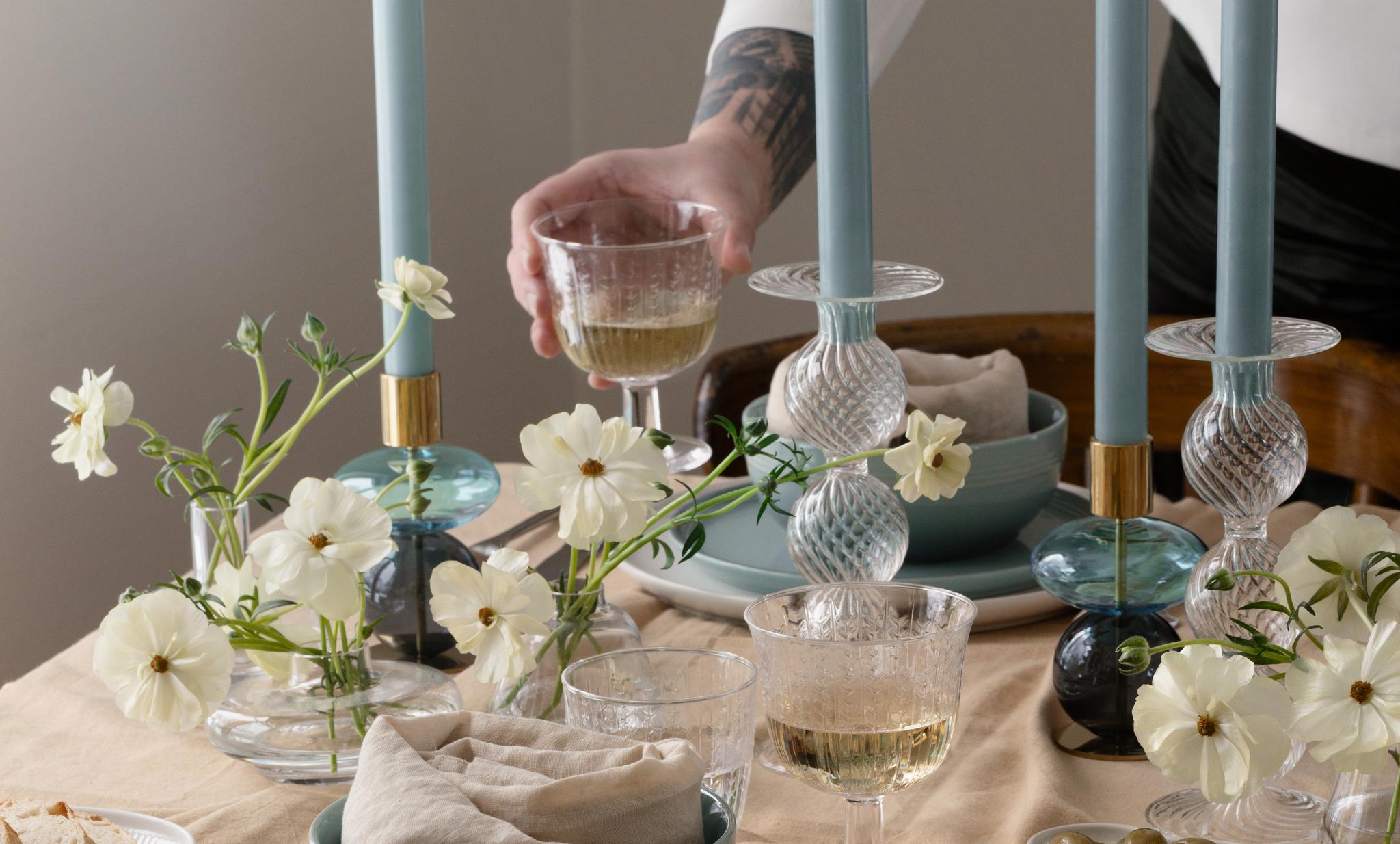 Tatuoitu käsivarsi asettaa viinilasin ruokapöydälle, jolla on sinisiä kynttilöitä, valkoisia kukkia, sinivihreät ja valkoiset astiat, leipää ja oliiveja vaaleanruskealla liinalla.