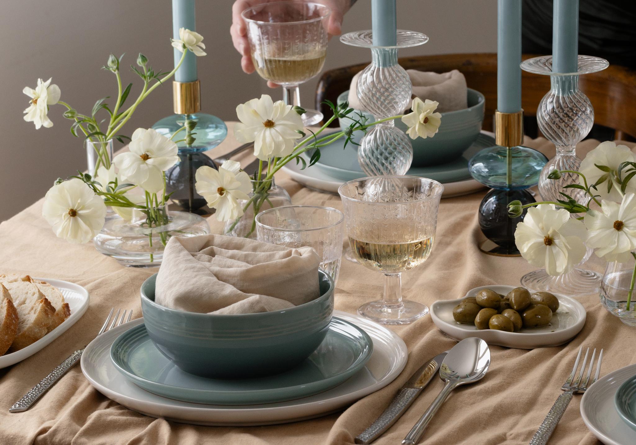 En hand håller ett vinglas bredvid ett bord dekorerat med blå koniska ljus i dekorativa hållare och vita blommor.