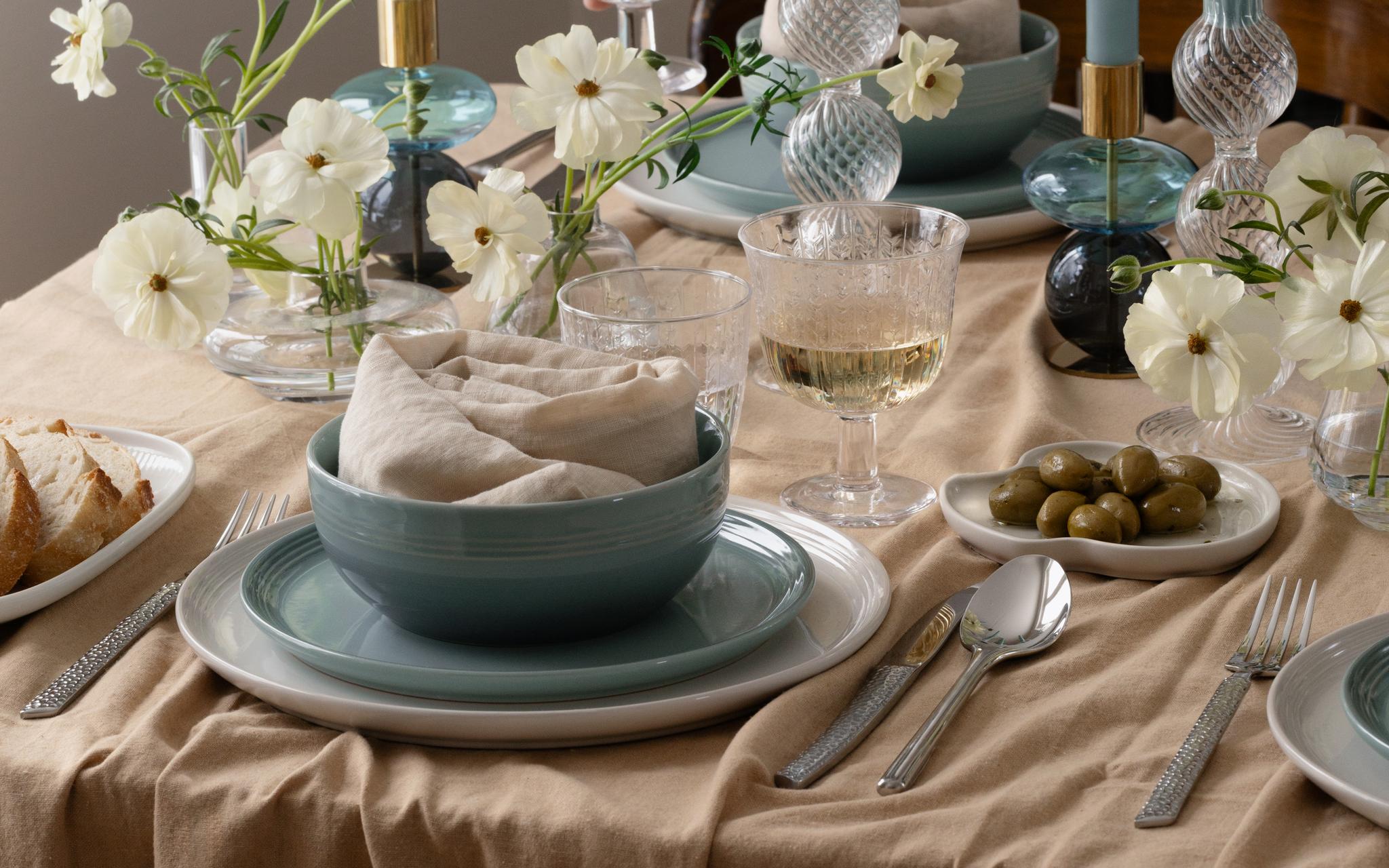 Henkilön tatuoitu käsivarsi kurottaa kohti lasillista valkoviiniä ruokapöydällä, jonka päällä on vaaleansinisiä astioita, valkoisia kukkia ja sytyttämättömiä sinisiä lamppukynttilöitä.