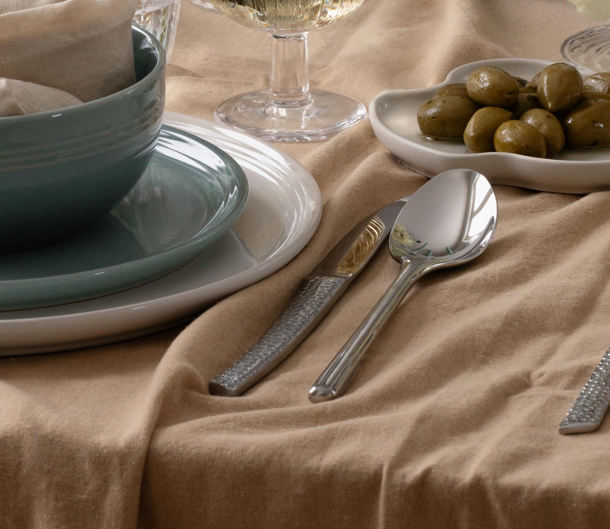 Henkilön käsi pitelee viinilasia ruokapöydällä, jolla on vaaleansinisiä kynttilöitä, valkoisia kukkia, sinivihreät astiat ja ruskea pöytäliina.